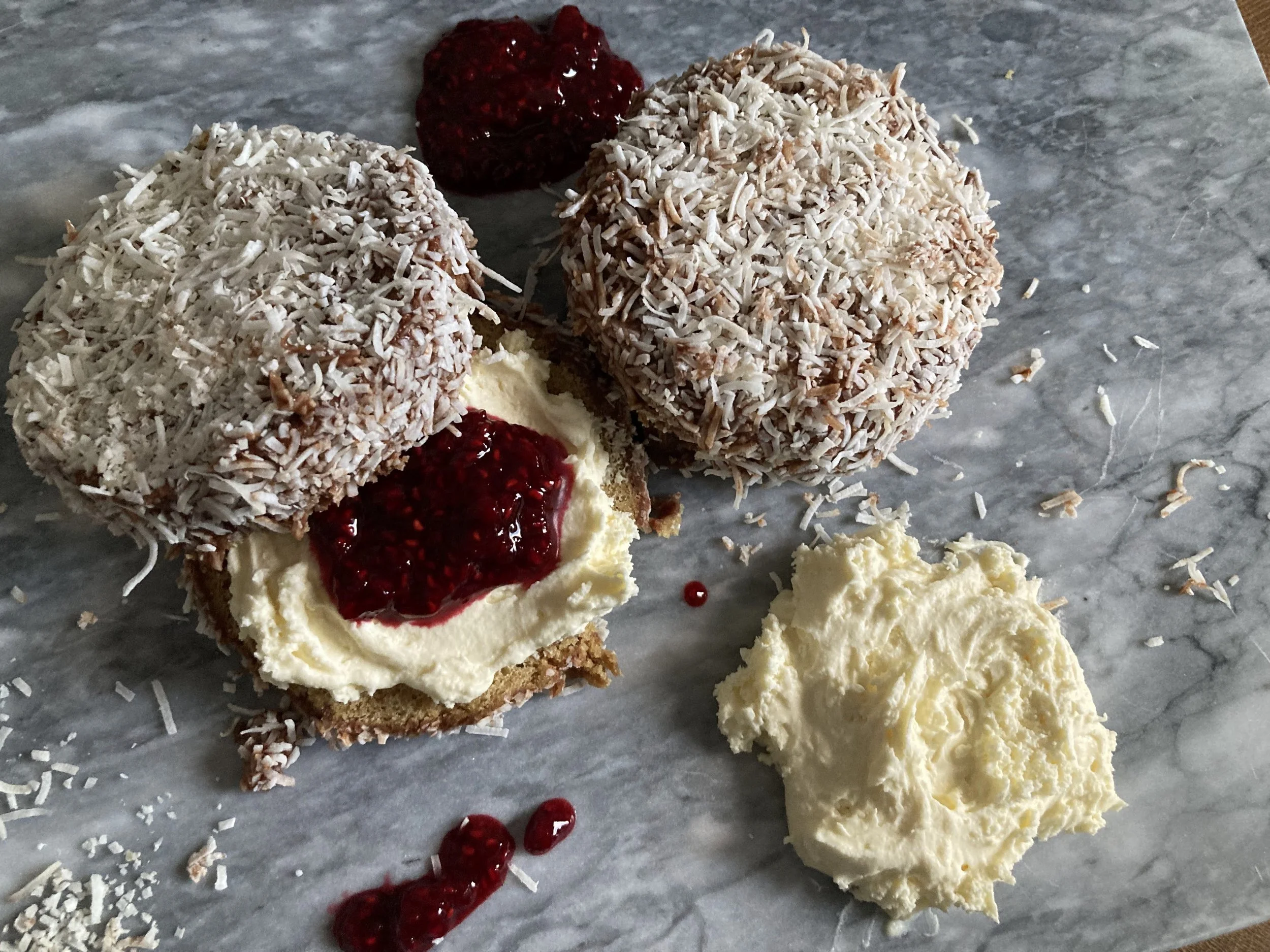 Two lamington cookies with shredded coconut, one cut open showing sponge cake, cream, and raspberry jam filling, with a side of whipped cream and some raspberry jam on a gray marble surface.
