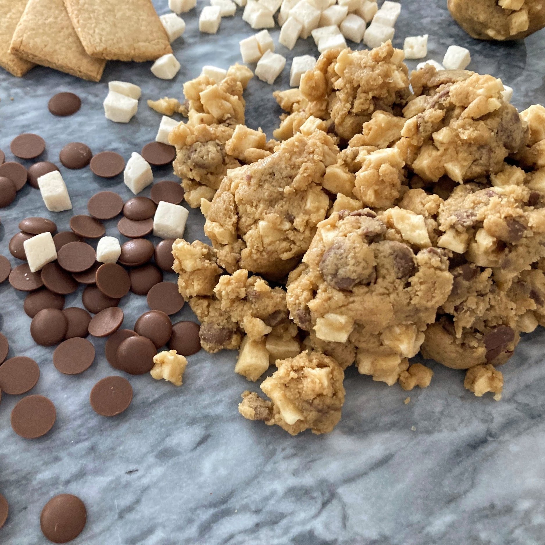 Chunks of cookie dough, mini chocolate chips, vanilla marshmallows, and graham crackers scattered on marble, resembling ingredients for s'mores.