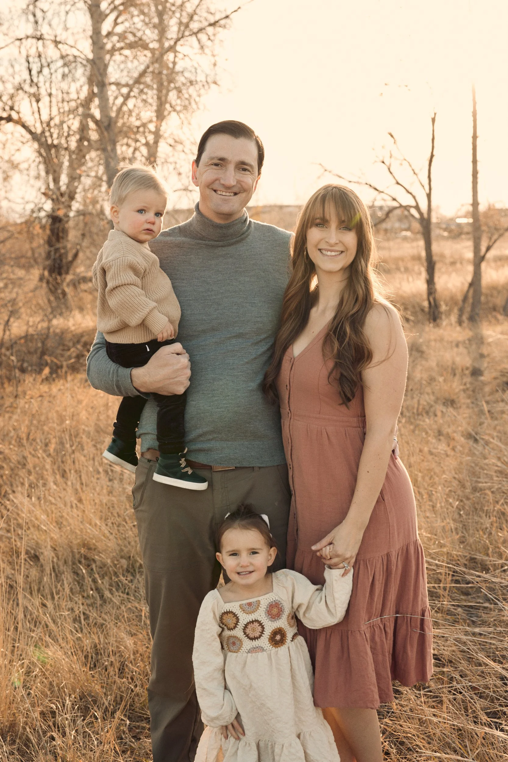 A family of four outdoors during sunset in a field with tall, dry grass and leafless trees, smiling at the camera.