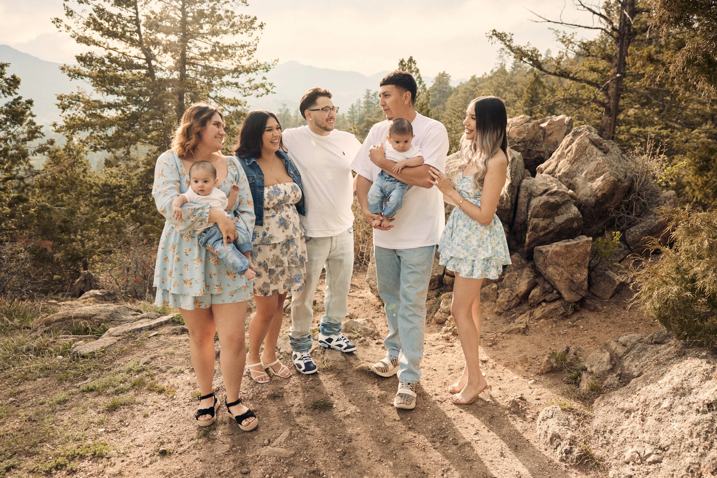 A group of six people, including children, standing outdoors in a rocky, wooded area with mountains in the background, smiling and chatting.