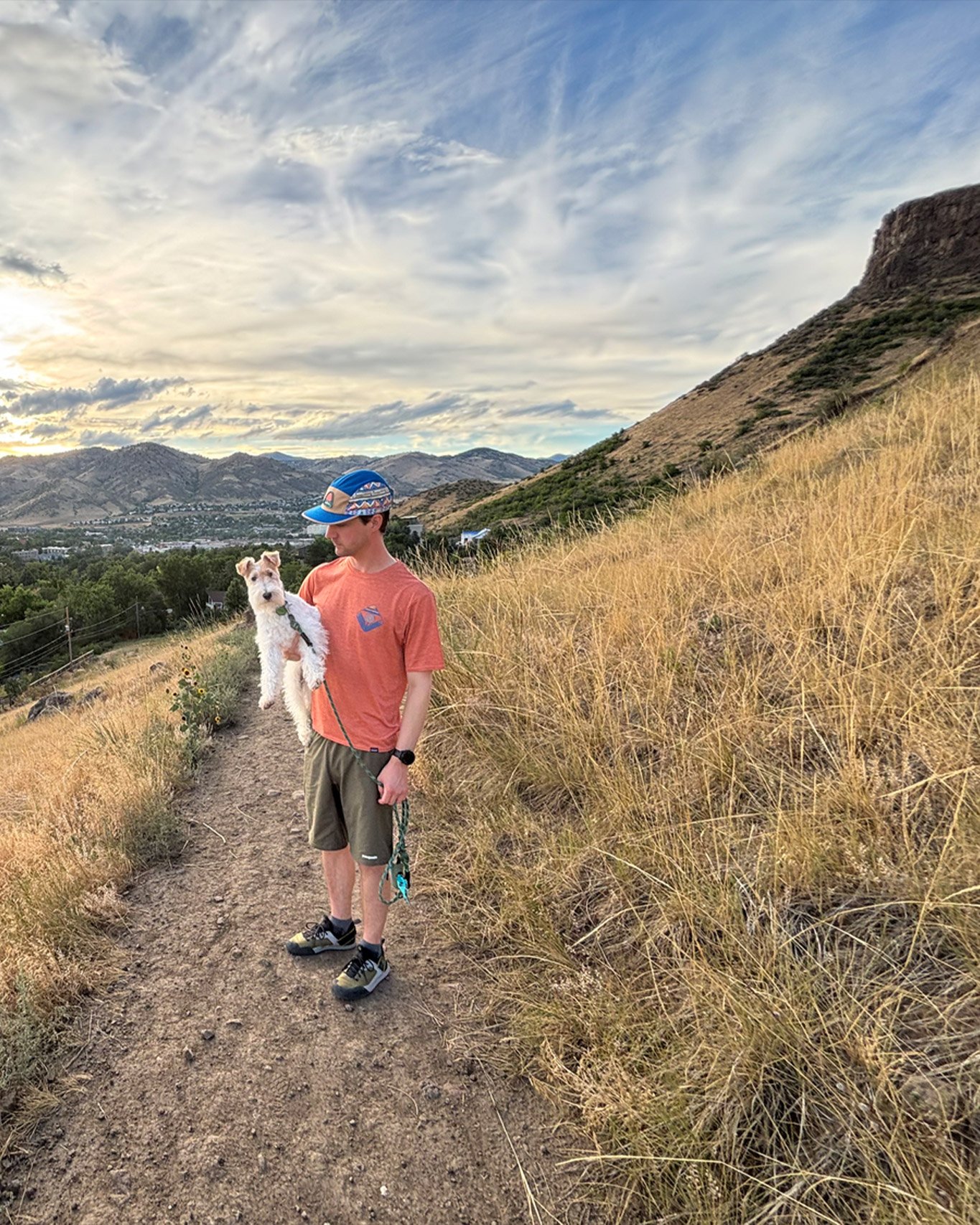 Person holding a dog on a hiking trail with mountainous landscape and cloudy sky in the background.