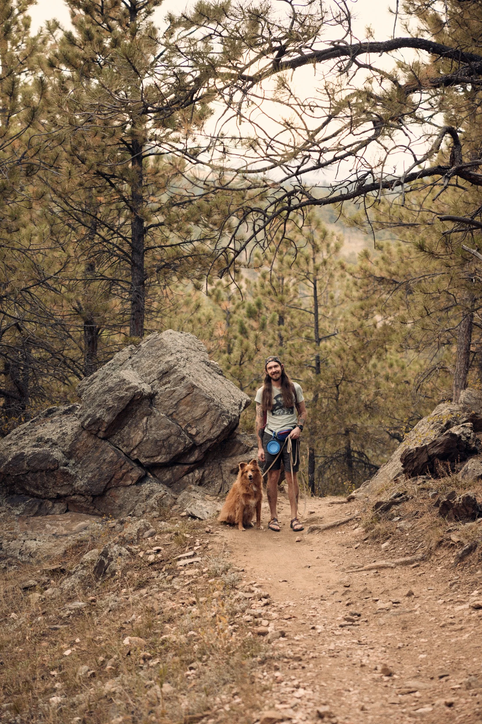 A man with long hair, beard, wearing a t-shirt, shorts, and sandals, standing next to a large, fluffy, tan dog outdoors on a trail surrounded by rocks, trees, and pine forest with mountains in the background.