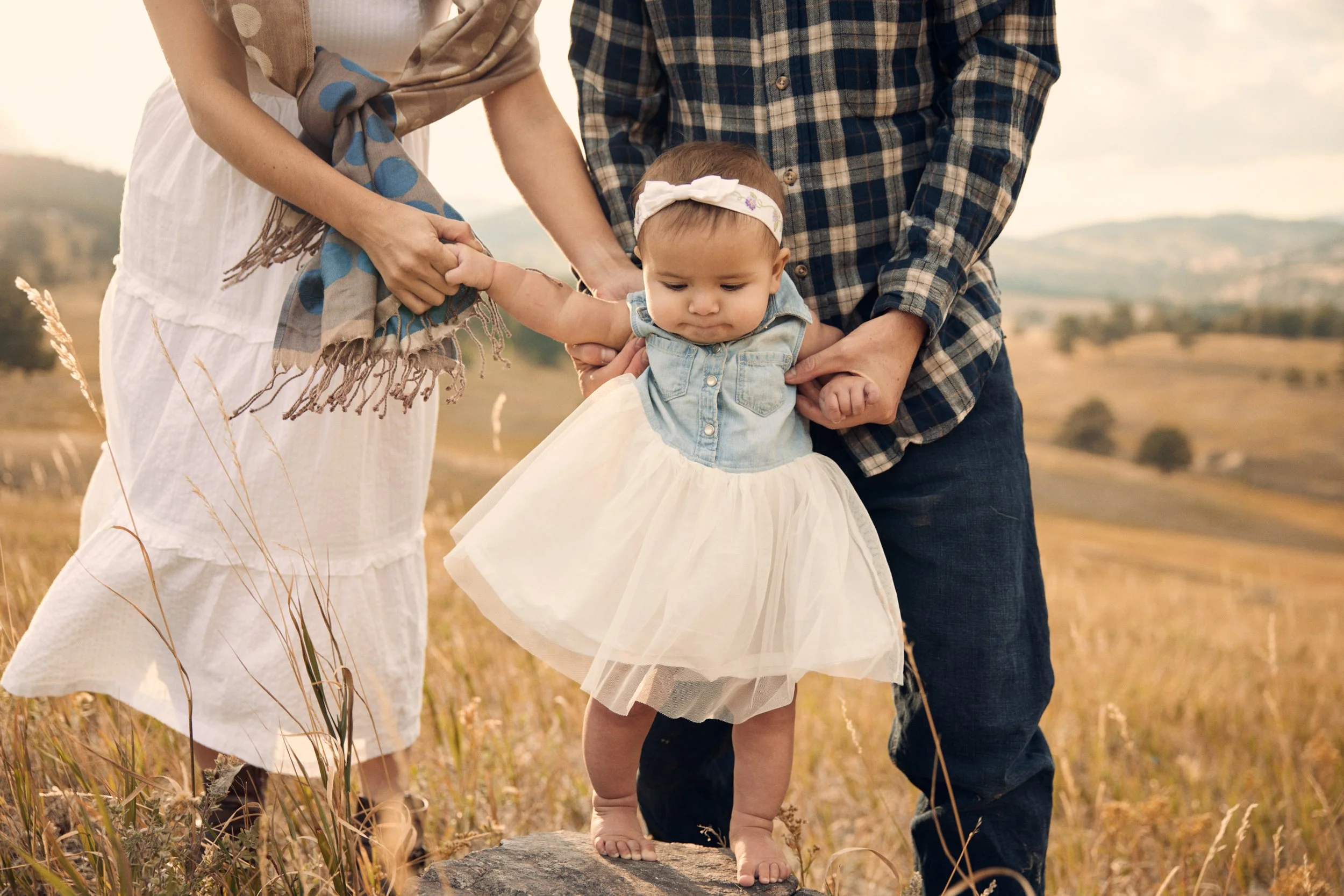 A young girl with a white headband and a white dress walking on rocks in a field, being supported by a woman and a man.