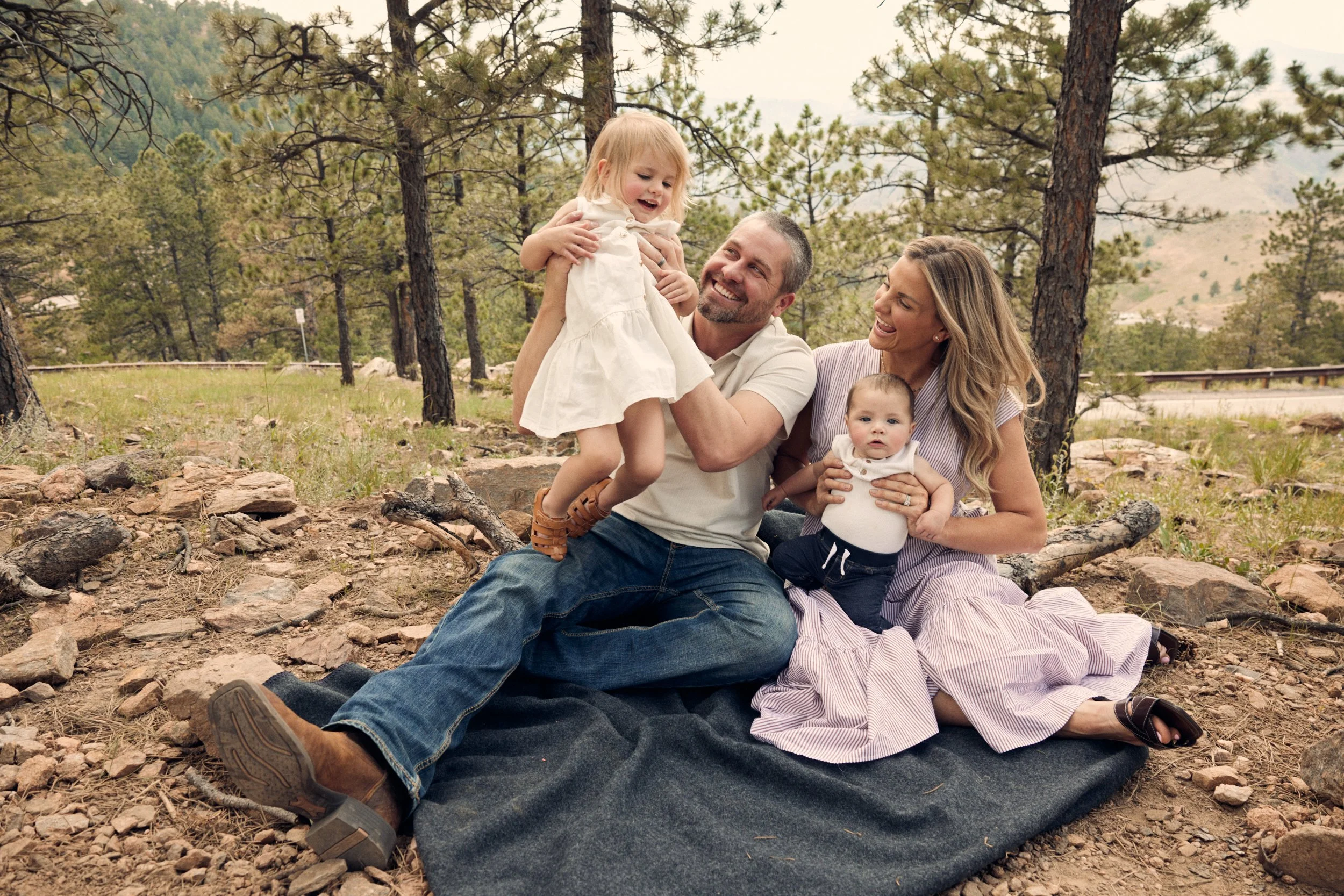 A family of four enjoying a moment outdoors in a forested area, with trees and mountains in the background. The father lifts his young daughter, while the mother holds their baby, and they all seem happy and engaged with each other.