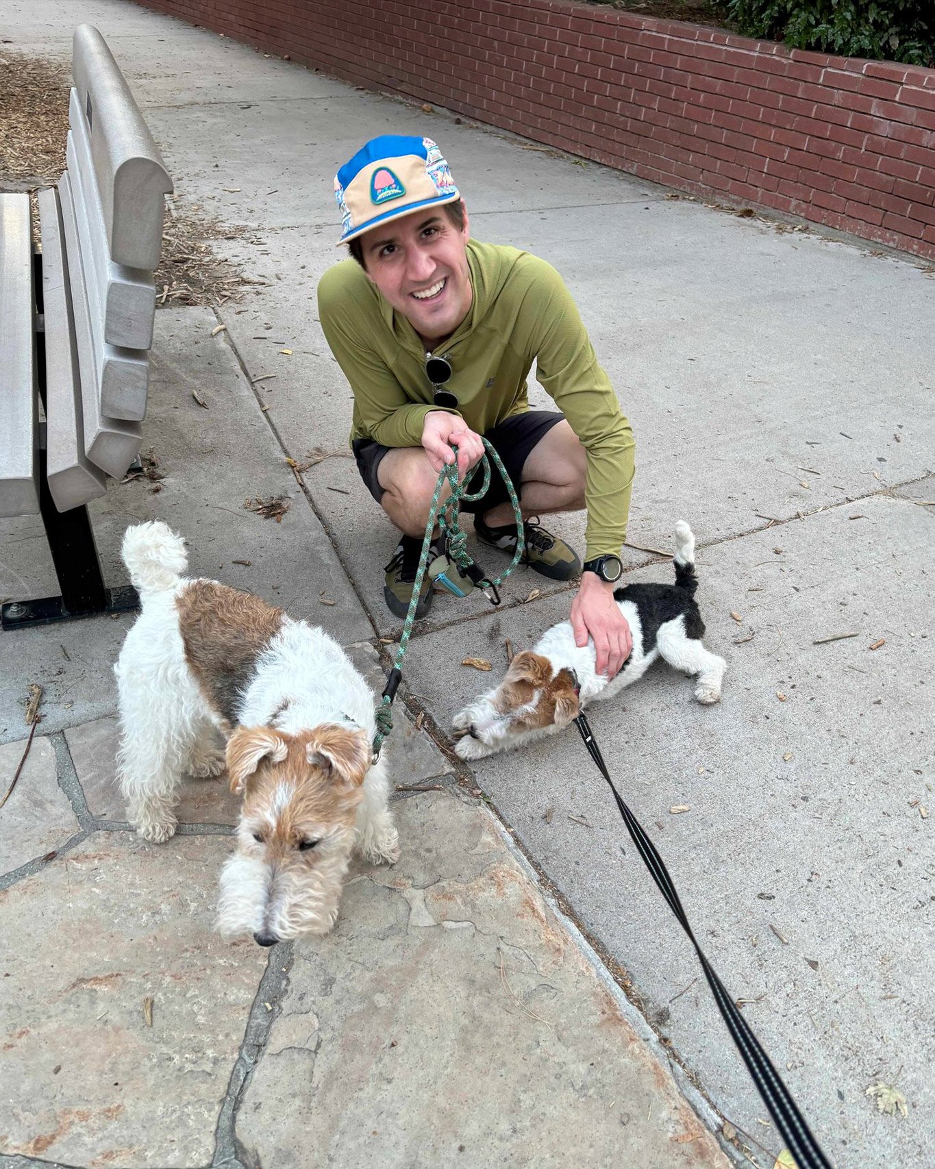 Person squatting on a sidewalk, smiling, holding leashes of two wire fox terriers, near a park bench and brick wall.