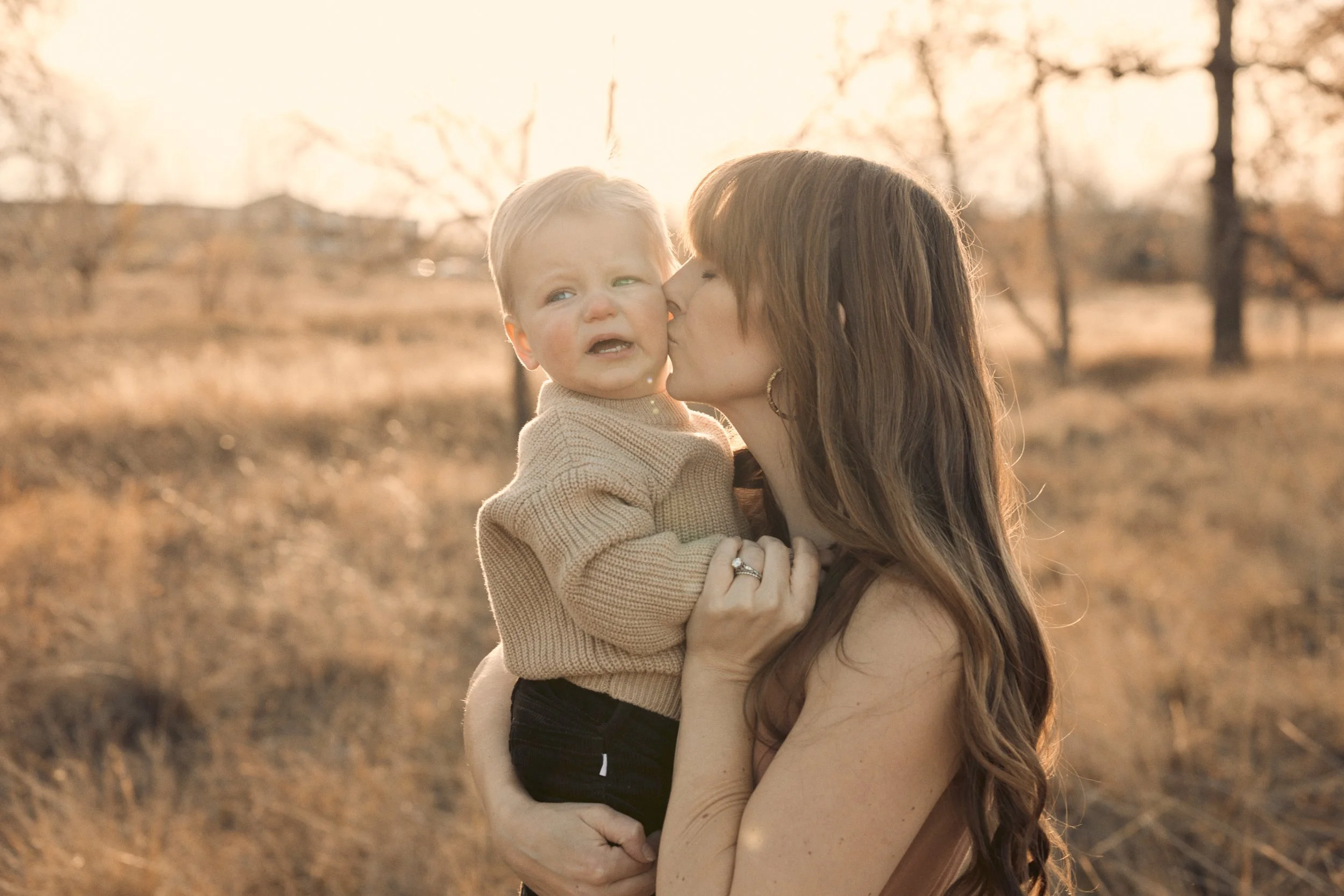 A woman with long brown hair holding a crying toddler son outdoors during sunset with trees and a field in the background.