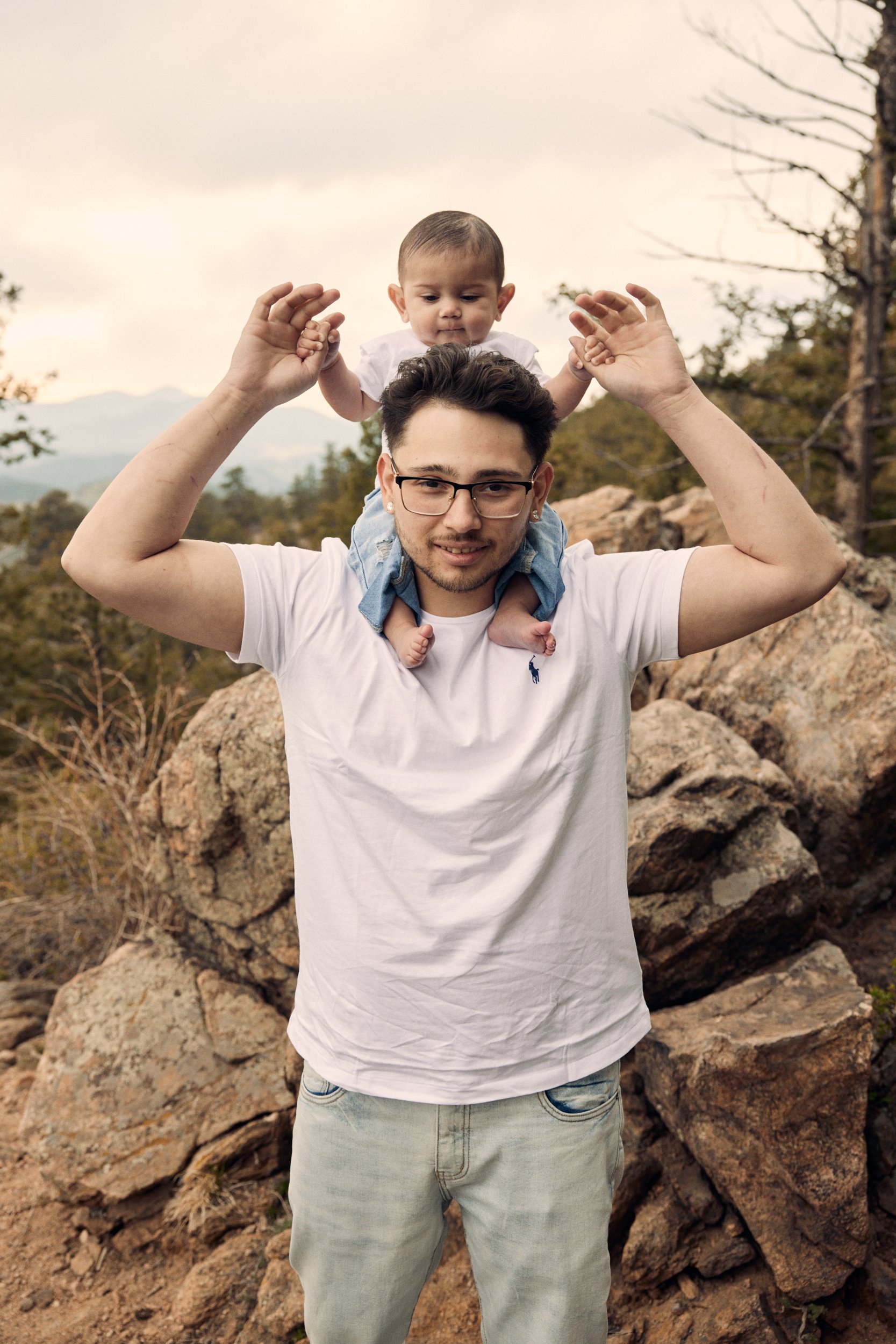 A man with glasses and a white t-shirt standing outdoors on rocky terrain, carrying a young child on his shoulders, holding the child's hands. The background features trees and mountains under a cloudy sky.
