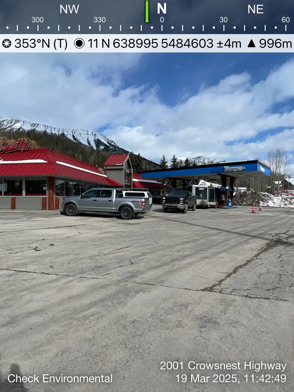 A parking lot in front of a restaurant and a gas station with snow-capped mountains in the background under a partly cloudy sky.