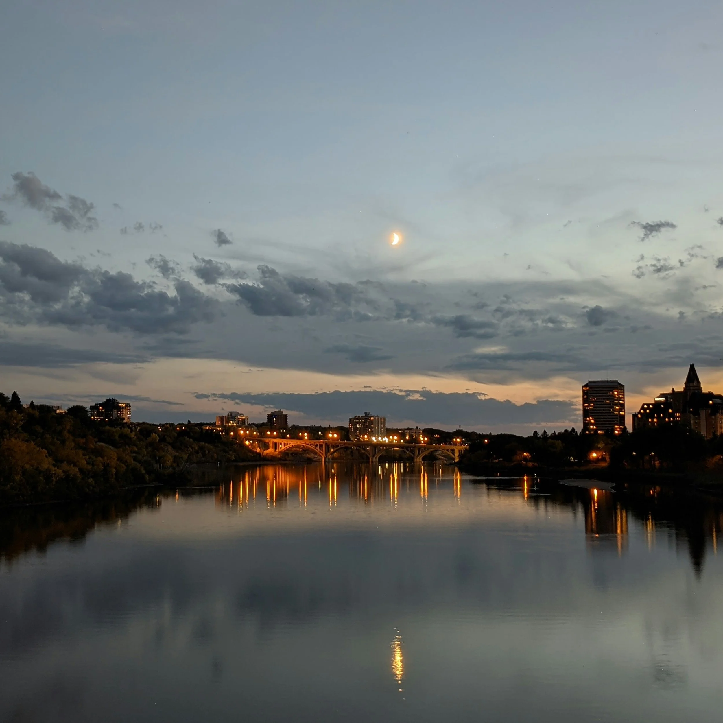 City skyline at dusk with illuminated buildings and their reflections on the calm river, under a sky with a crescent moon and scattered clouds.