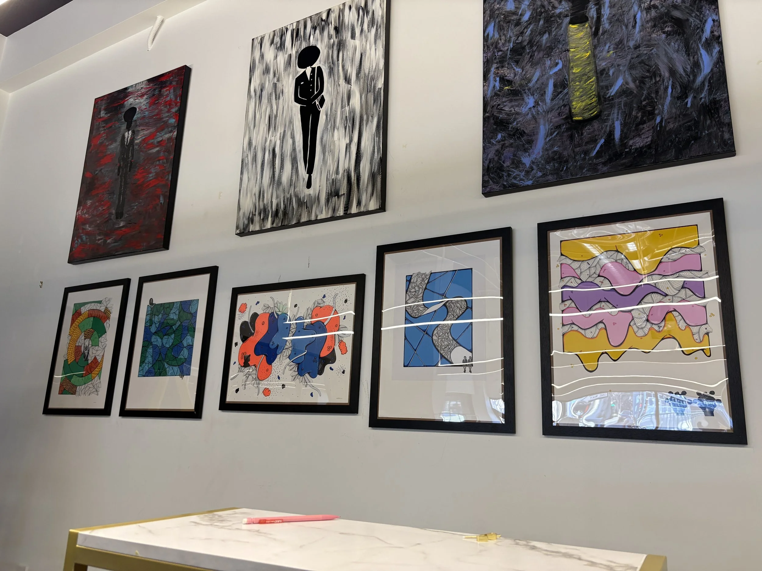 A photo of artwork on display In The Black Shop's wall. Pictured are three paintings by André Renay and five mixed media illustrations by Ms. Mac.