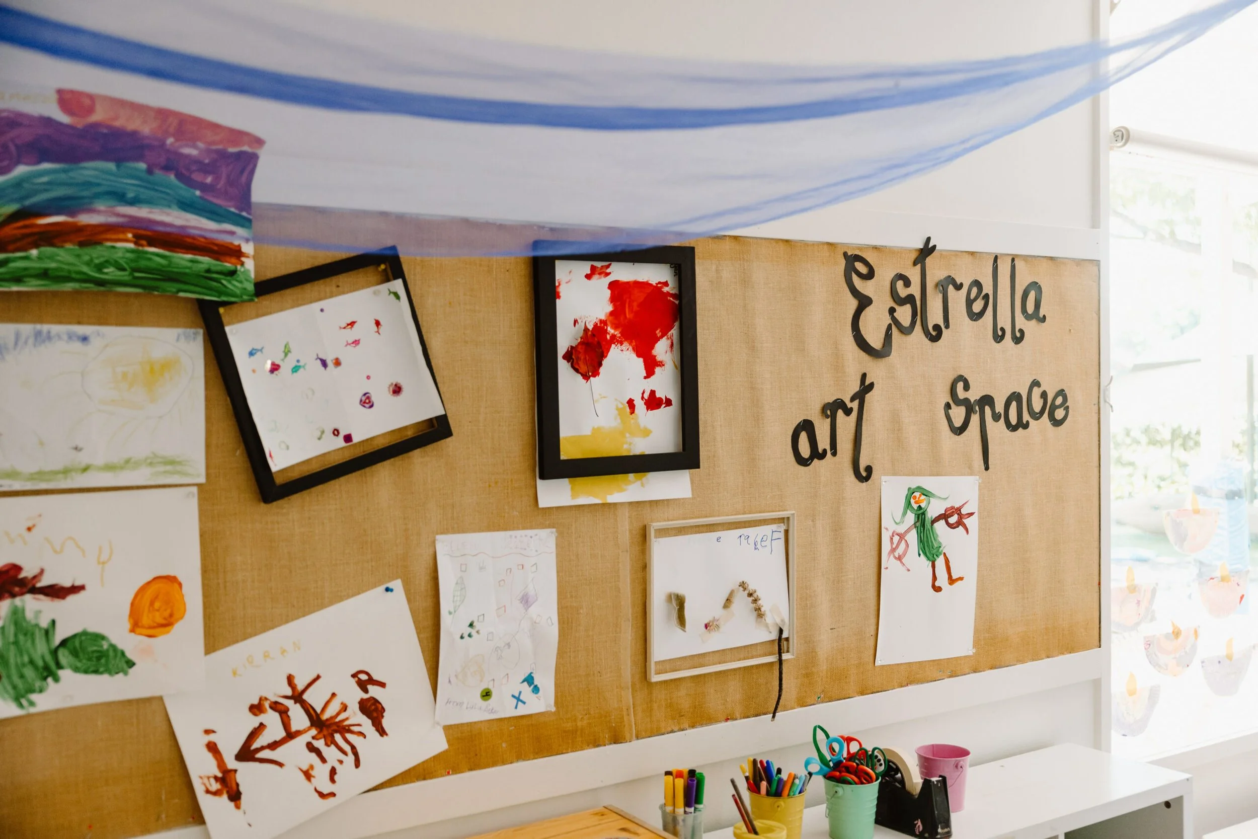 Children's artwork displayed on a tan bulletin board labeled 'Estrella art space' with various drawings and paintings, some in frames, in a classroom or art room setting.