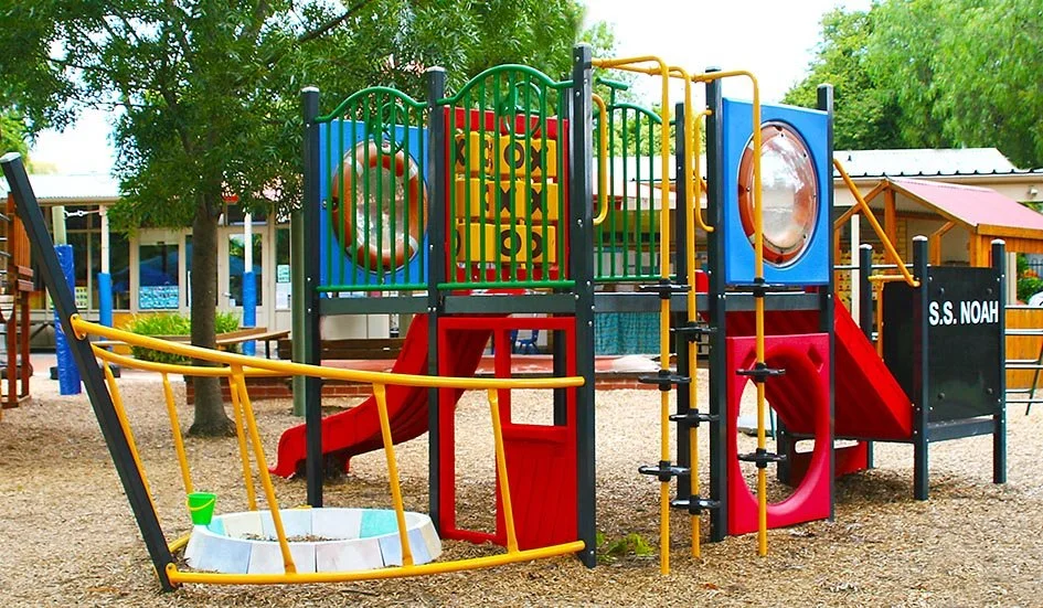 Colorful playground structure with slides, climbing bars, and a circular window, located outdoors with trees in the background.