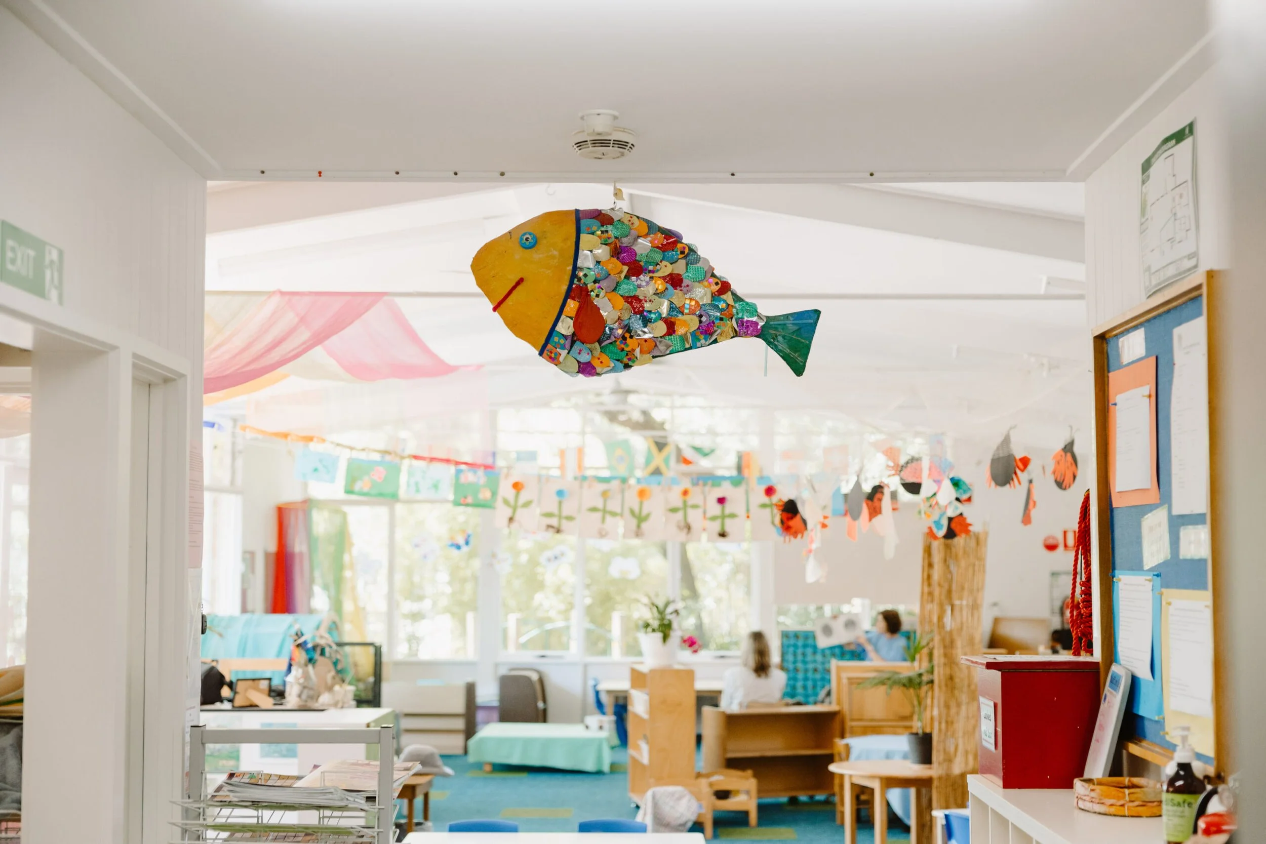 Colorful fish-shaped hanging decoration in a bright, child-friendly room with art displays, furniture, and children playing.