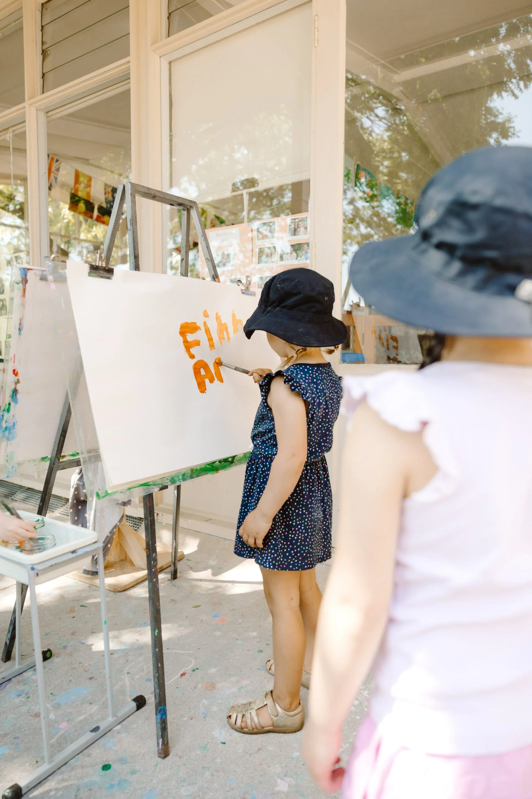 Two young girls are painting on a large white canvas outside, with the words "Film and" painted in orange. Both girls are wearing black wide-brimmed hats, one in a dark, polka-dotted dress and the other in a white top with pink bottoms.