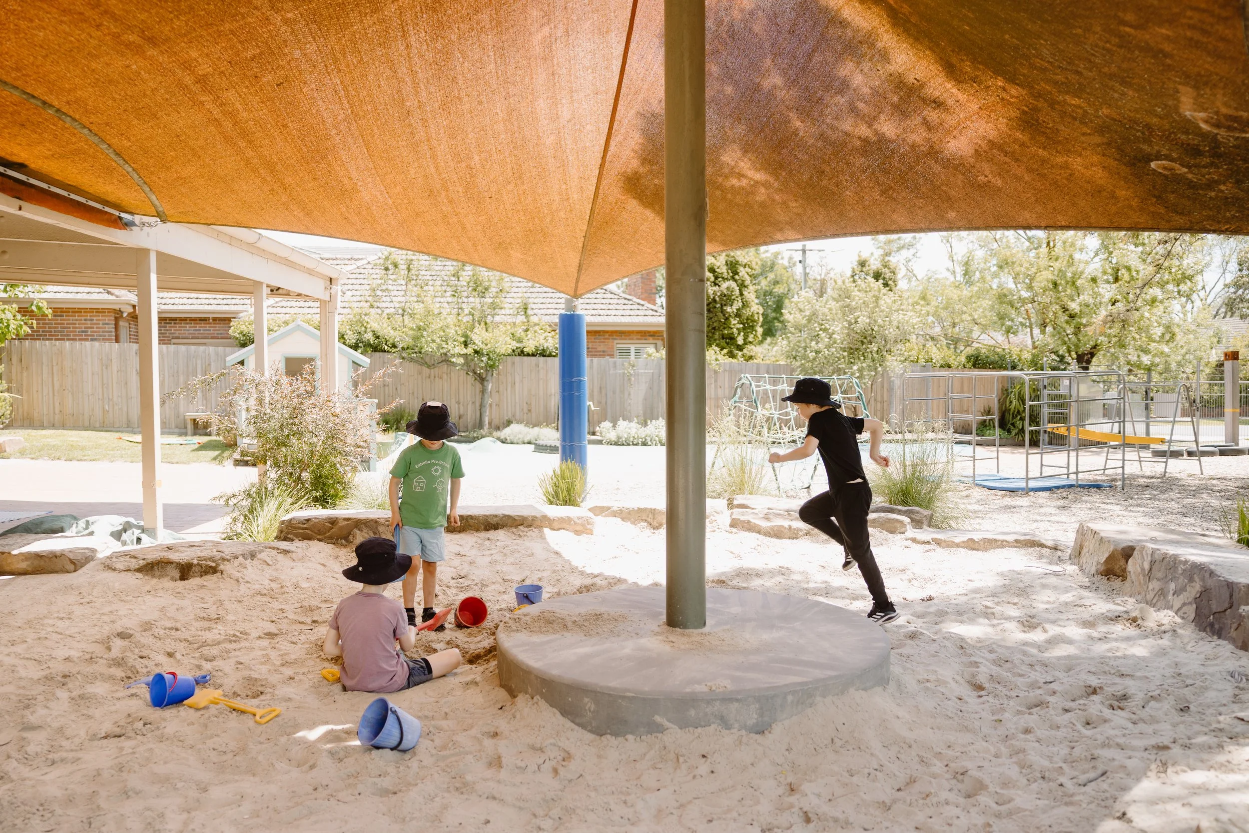 Children playing in a sandbox area at a playground, with a shade sail overhead and playground equipment in the background.