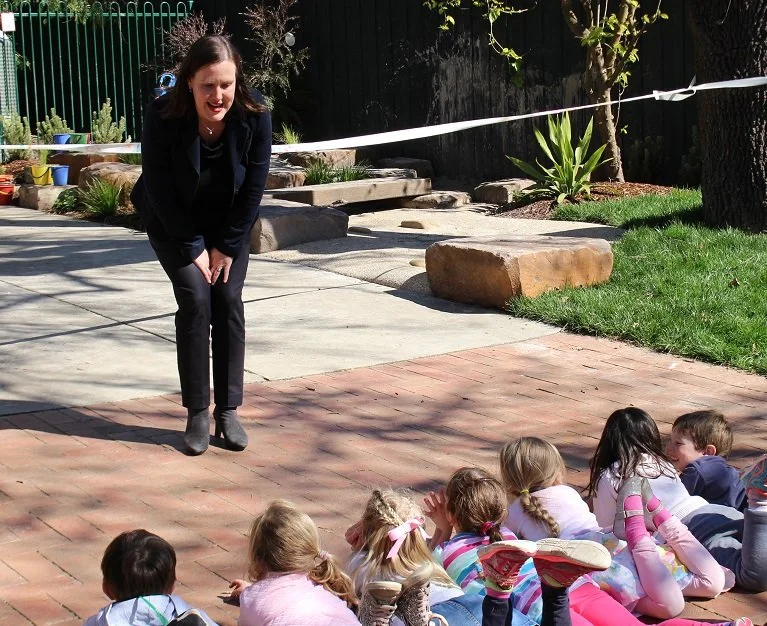 The Estrella Director standing outside, bending slightly forward, addressing a group of children who are lying on the ground and listening attentively.