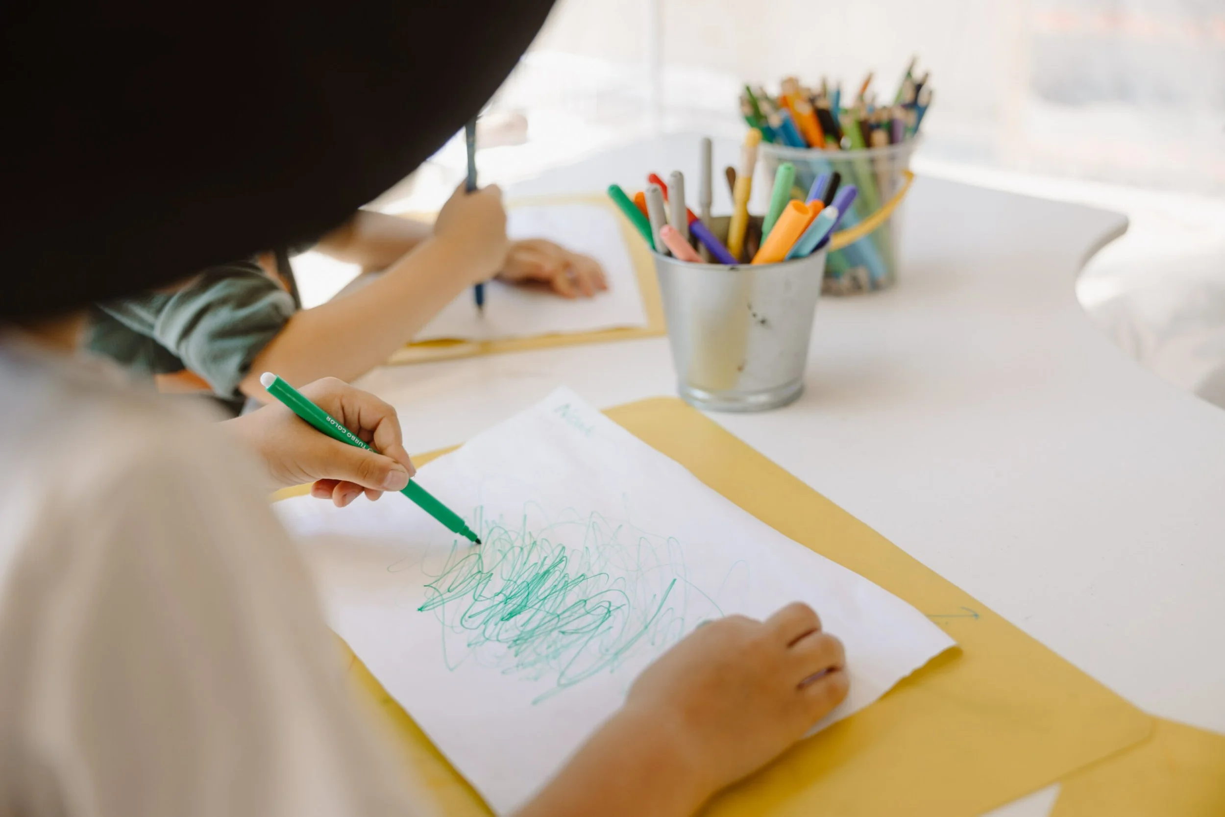 Child drawing with green marker on white paper, with colorful markers in containers on white table