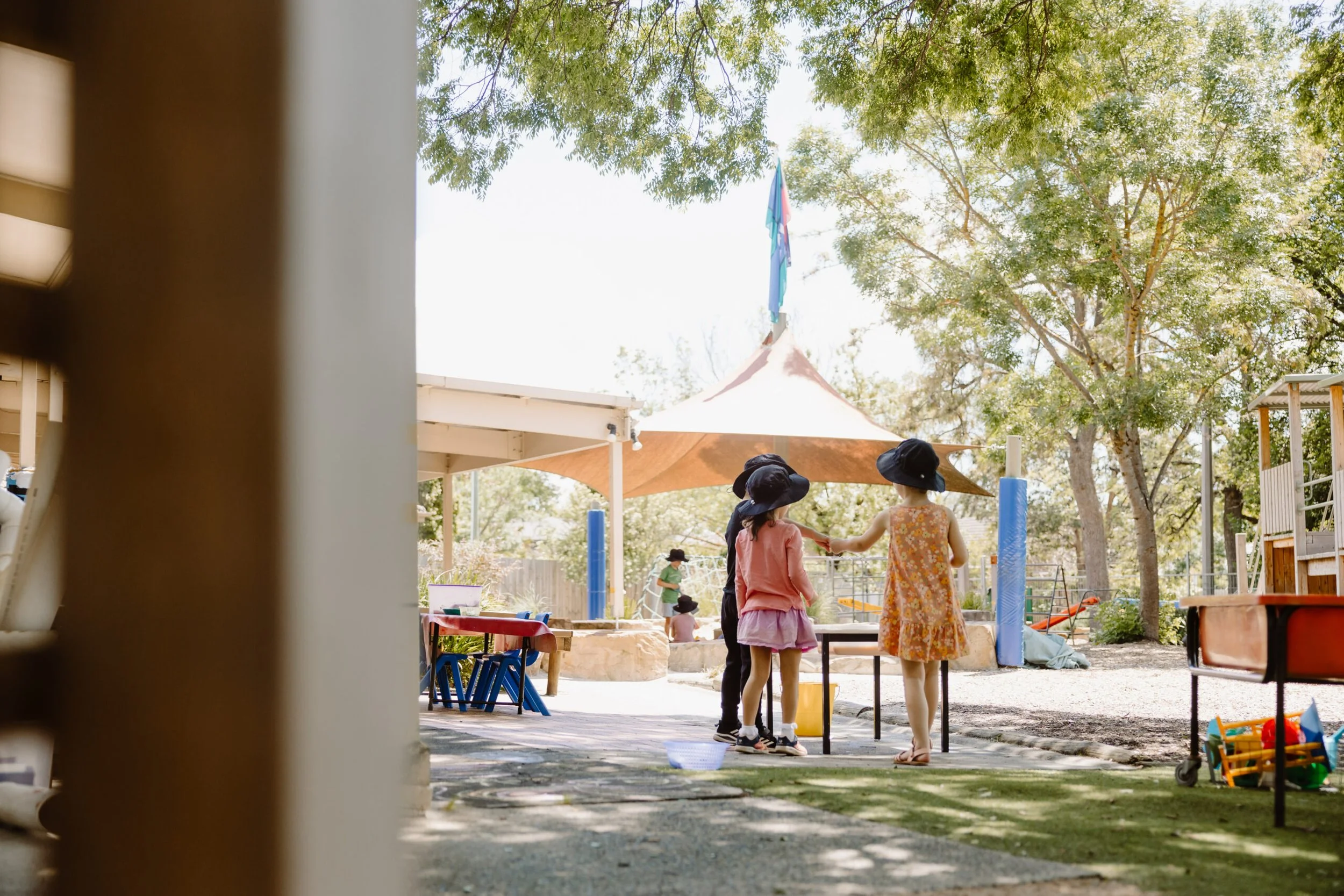 Children wearing wide-brimmed hats playing outside at a playground, with trees and a canopy in the background.