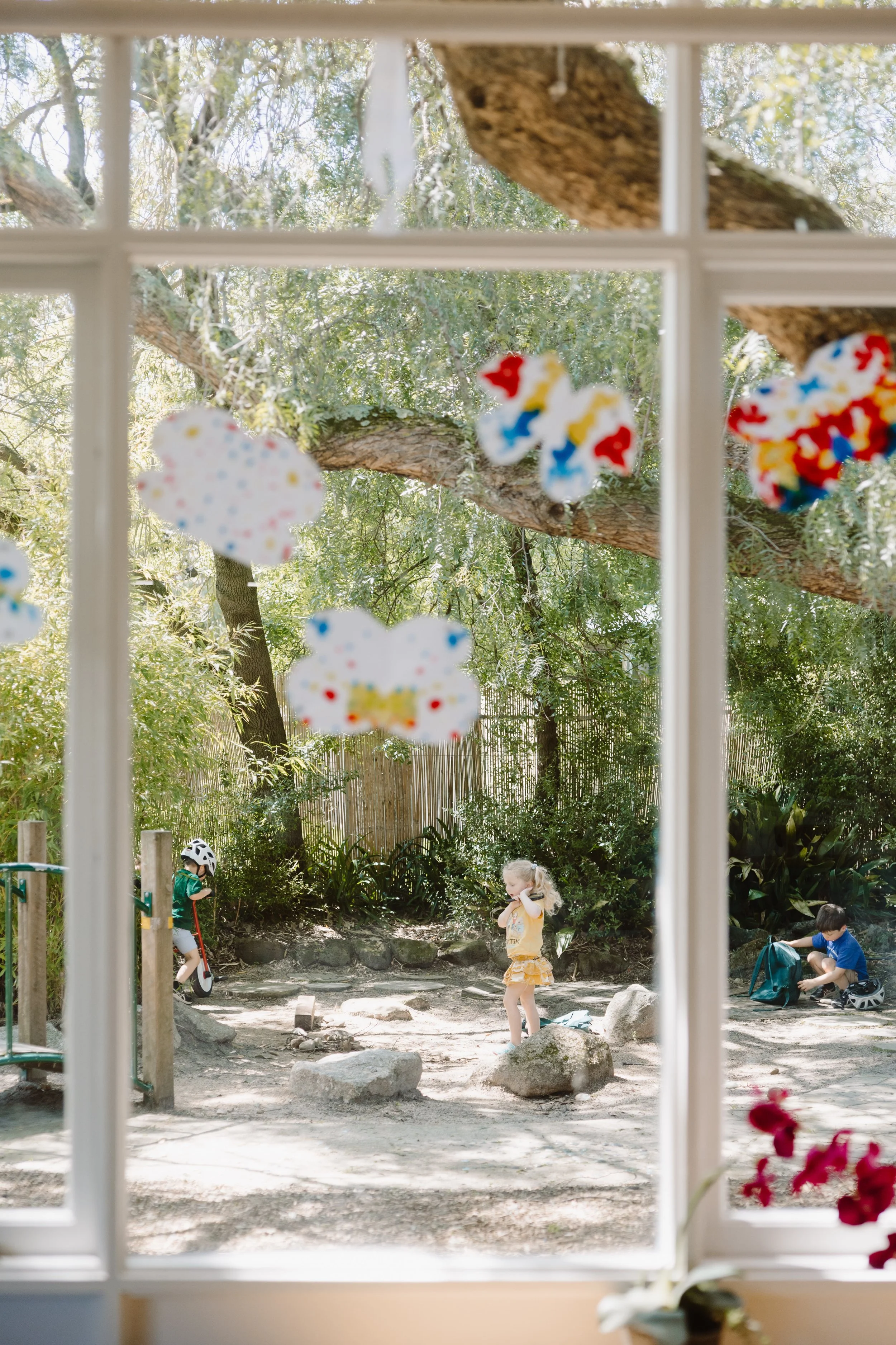 View through a window showing a small playground with children playing outside, surrounded by trees and greenery, with colorful decorations hanging in the foreground.