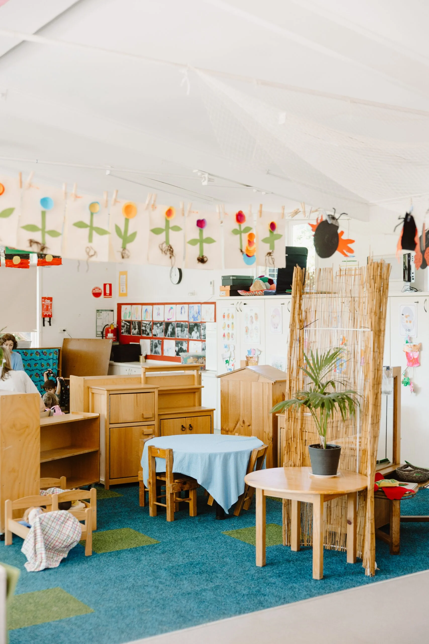 Colorful classroom decorated with paper flowers hanging from a string and various children's artwork on the walls. Child-sized furniture, including a round table with chairs, is arranged on a green and blue carpet. A potted plant is on a small wooden table in the foreground.