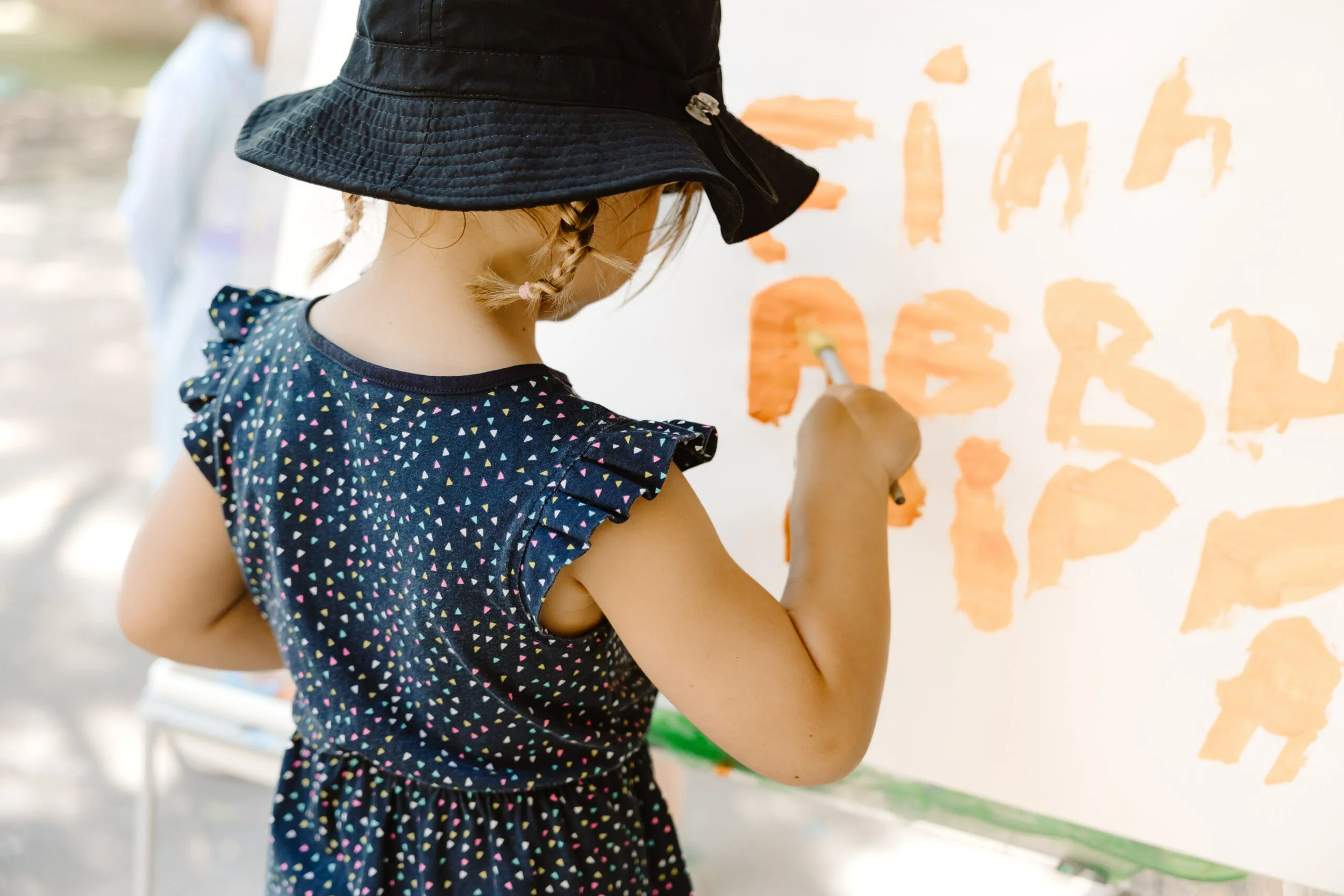 A young girl wearing a black bucket hat and a navy blue dress with multicolored dots, painting orange letters on a white surface.