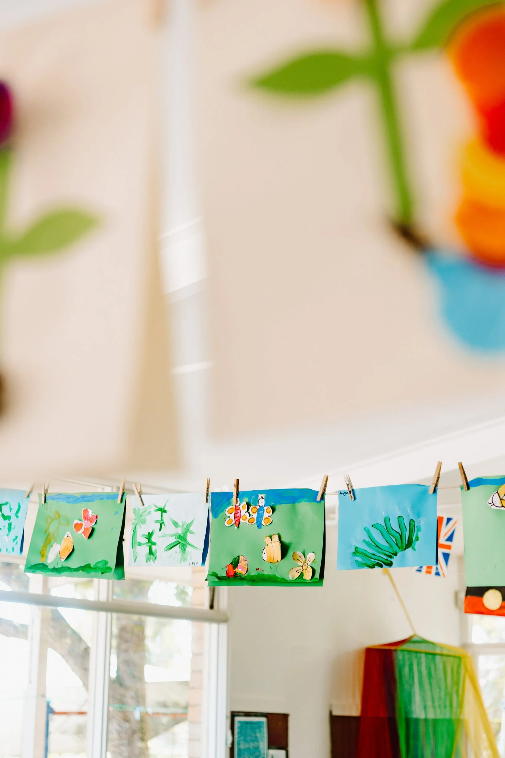 Colorful children's artwork hanging on a clothesline with clothespins in a classroom or art space, featuring butterflies, flowers, and nature themes.