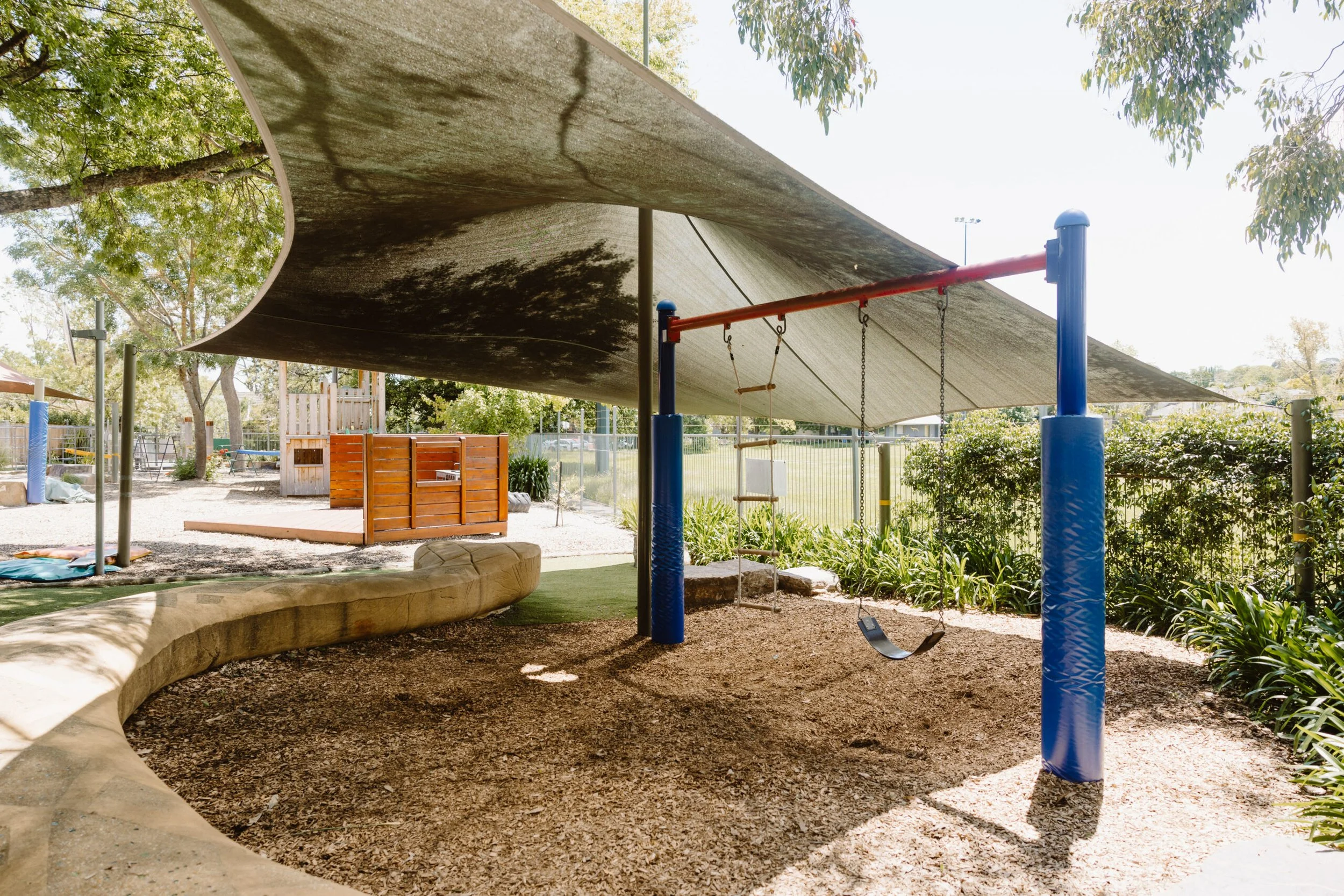 Empty playground area with a swing and shade sail overhead, surrounded by trees and greenery.