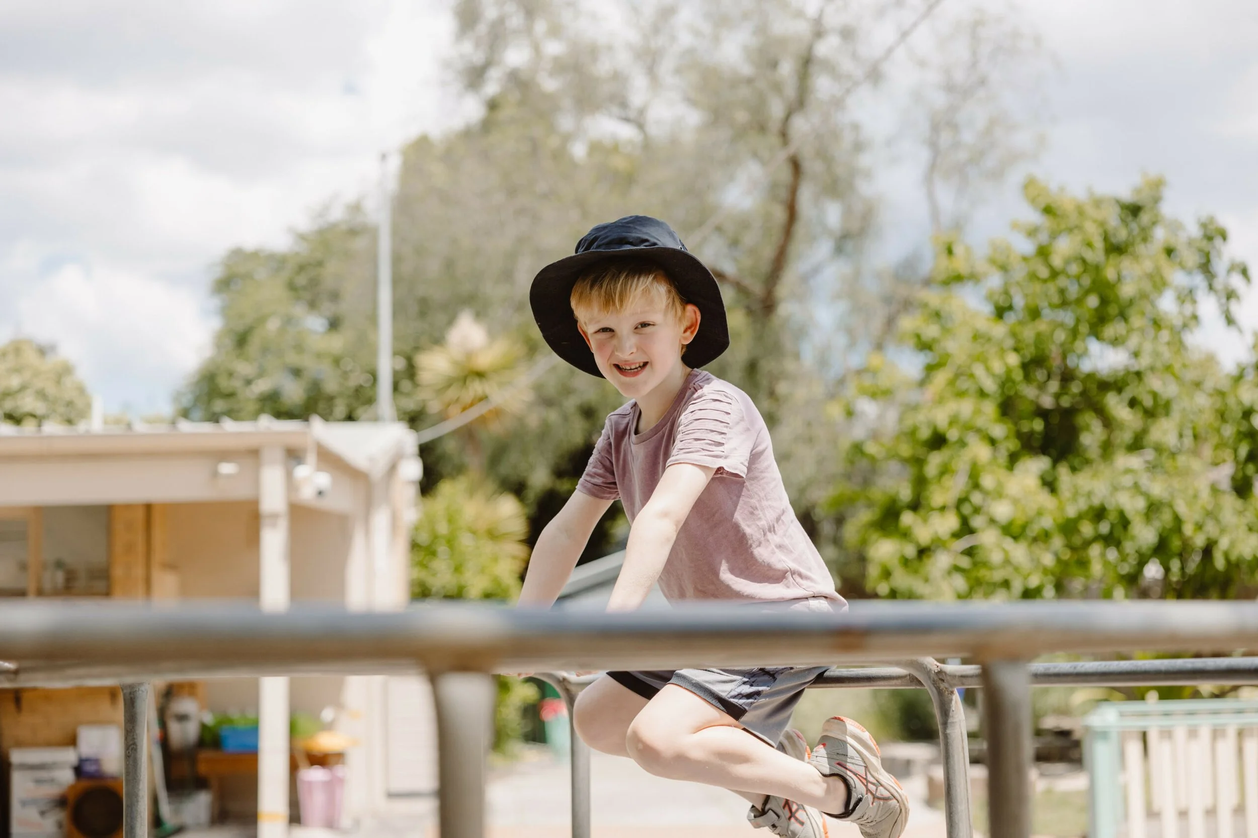 Young boy with a black hat playing on a jungle gym outdoors, smiling, with trees and a partly cloudy sky in the background.