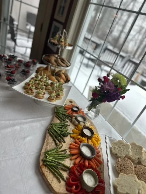An assortment of snacks and appetizers on a table, including sliced vegetables, crackers, and small bowls of dip, with a flower arrangement in a vase in the background near a window.