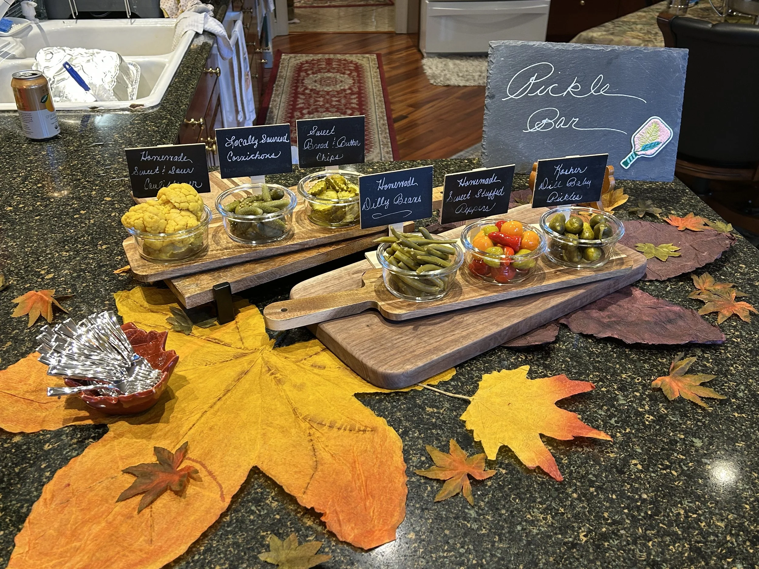 A fall-themed pickles bar with small bowls of homemade sweet and sour cucumbers, locally sourced cornichons, sweet bread and butter chips, homemade dill beans, sweet stuffed peppers, and fresh dill baby pickles on wooden trays. The bar is decorated with artificial colorful autumn leaves on a dark granite countertop, with a chalkboard sign reading 'PICKLE BAR' and an illustration of a pickle.