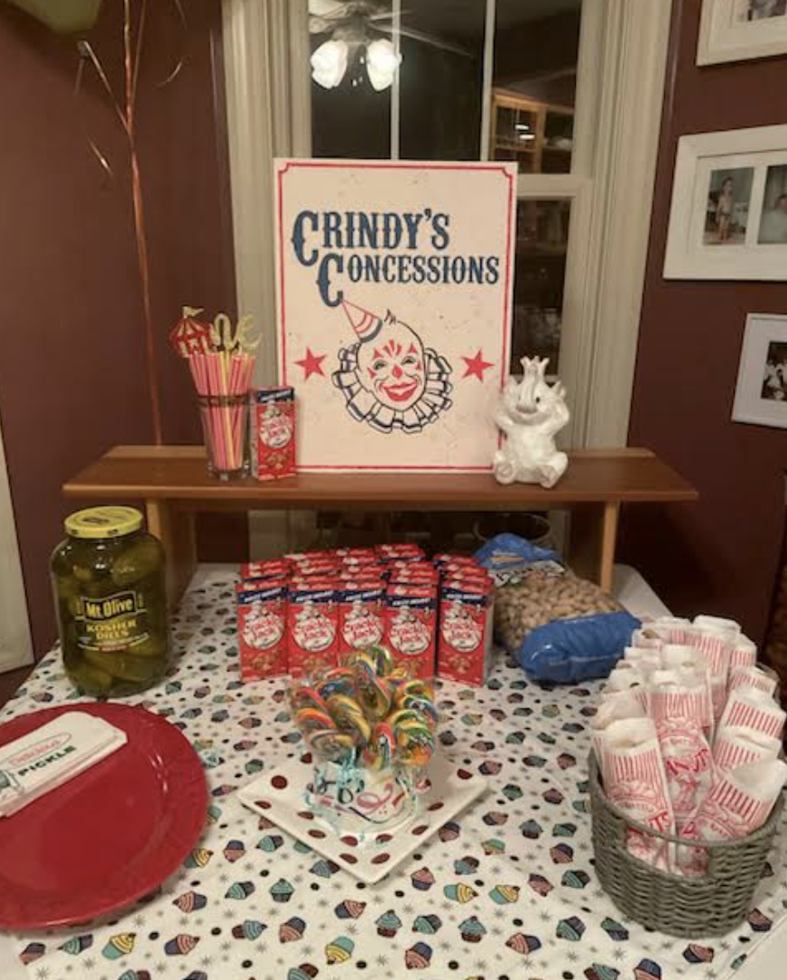 Table with snacks and concessions for a clown-themed event called 'Crindy's Concessions,' featuring a sign with a clown face, popcorn bags, lollipops, pickles, and a clown figurine.