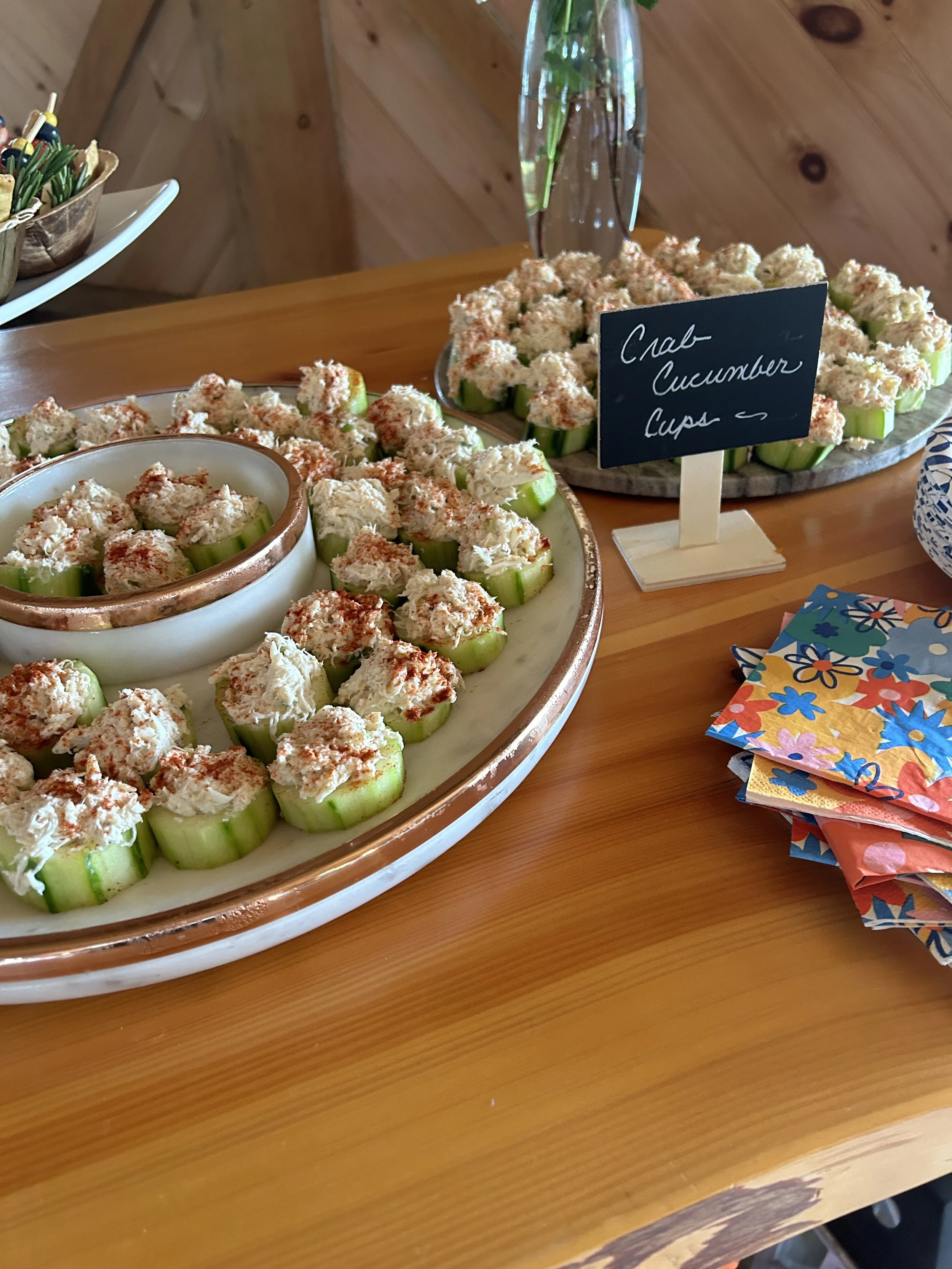 Cucumber cups filled with crab salad on a large serving platter, labeled as Crab Cucumber Cups, with colorful napkins beside them on a wooden table.
