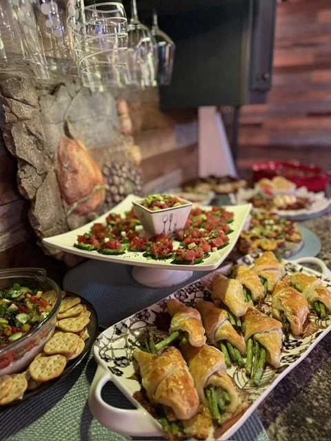 A variety of appetizers on a buffet table, including bacon-wrapped jalapeño poppers, a vegetable and dip platter, and a tomato and basil salad, set against a rustic stone wall and wooden backdrop.