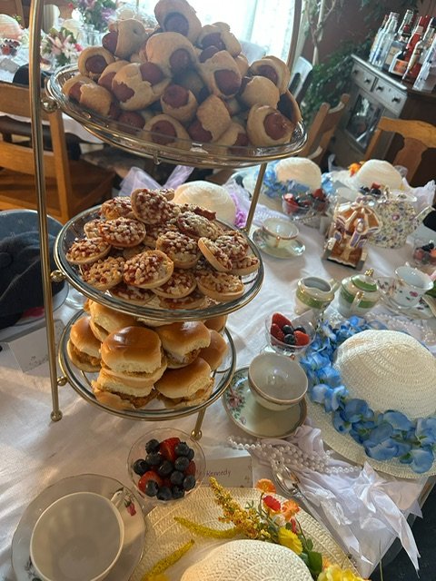 Three-tiered stand with assorted cookies and small sandwiches on a dining table, decorated with tea sets, fruit, and floral arrangements for a tea party.