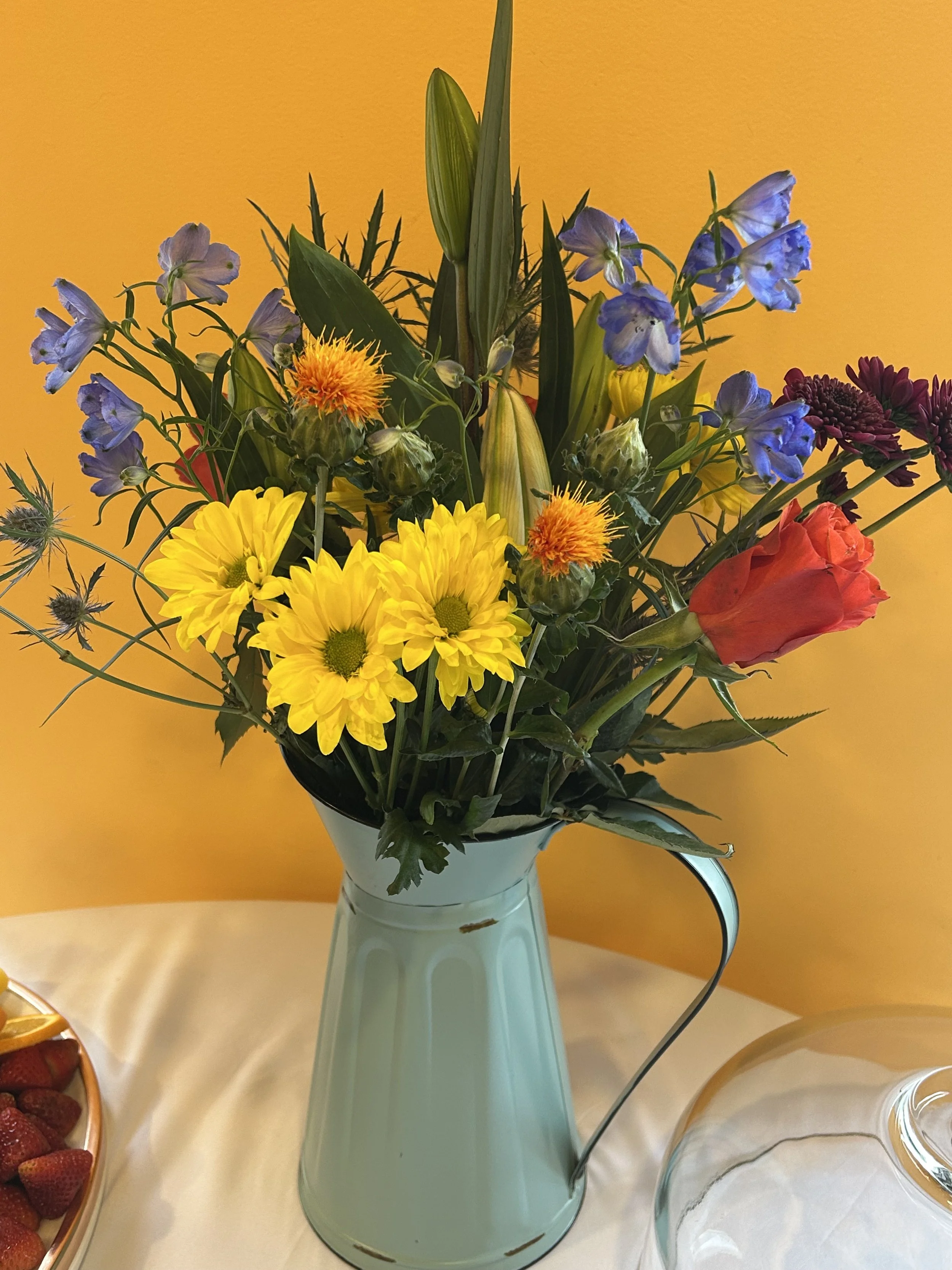 Bouquet of colorful flowers in a blue vase on a white table with a yellow wall background.