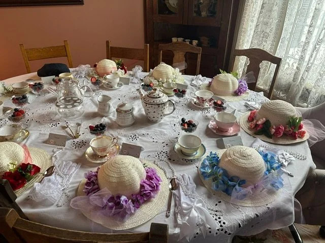 A round dining table decorated with lace tablecloths, featuring several straw hats adorned with fabric and artificial flowers, surrounded by teacups, teapots, bowls with berries, and small name cards, set in a room with floral curtains and wooden chairs.