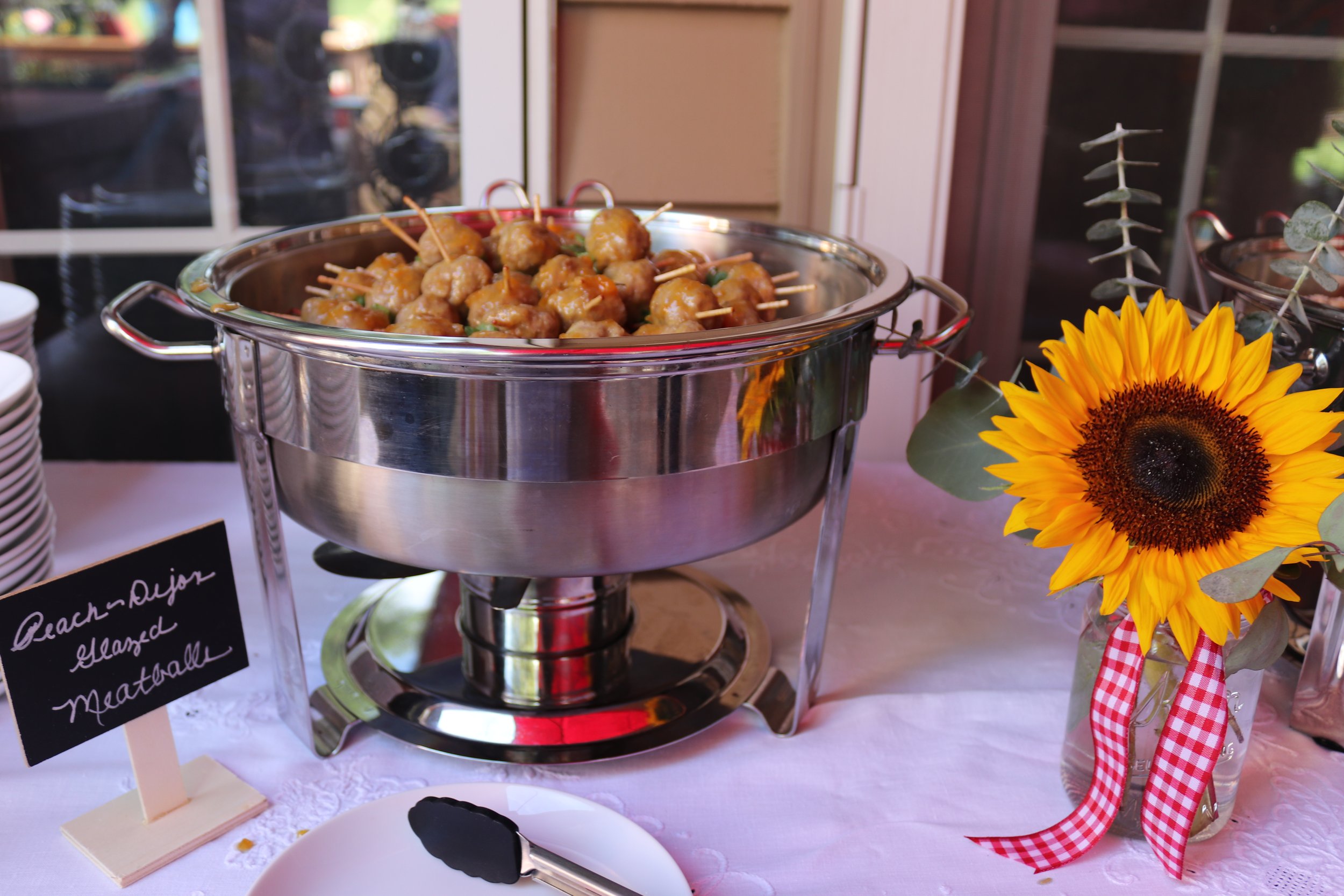 Chafing dish filled with meatballs and skewers, with a sunflower in a vase decorated with a red gingham ribbon next to it.