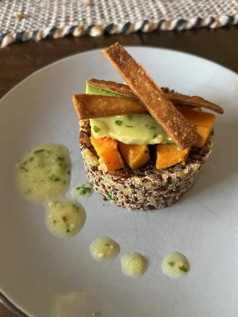 A plated dish featuring a quinoa cup filled with roasted sweet potatoes, avocado slices, and crispy breadstick garnishes, with a light green herb sauce sauce dotted on the side.