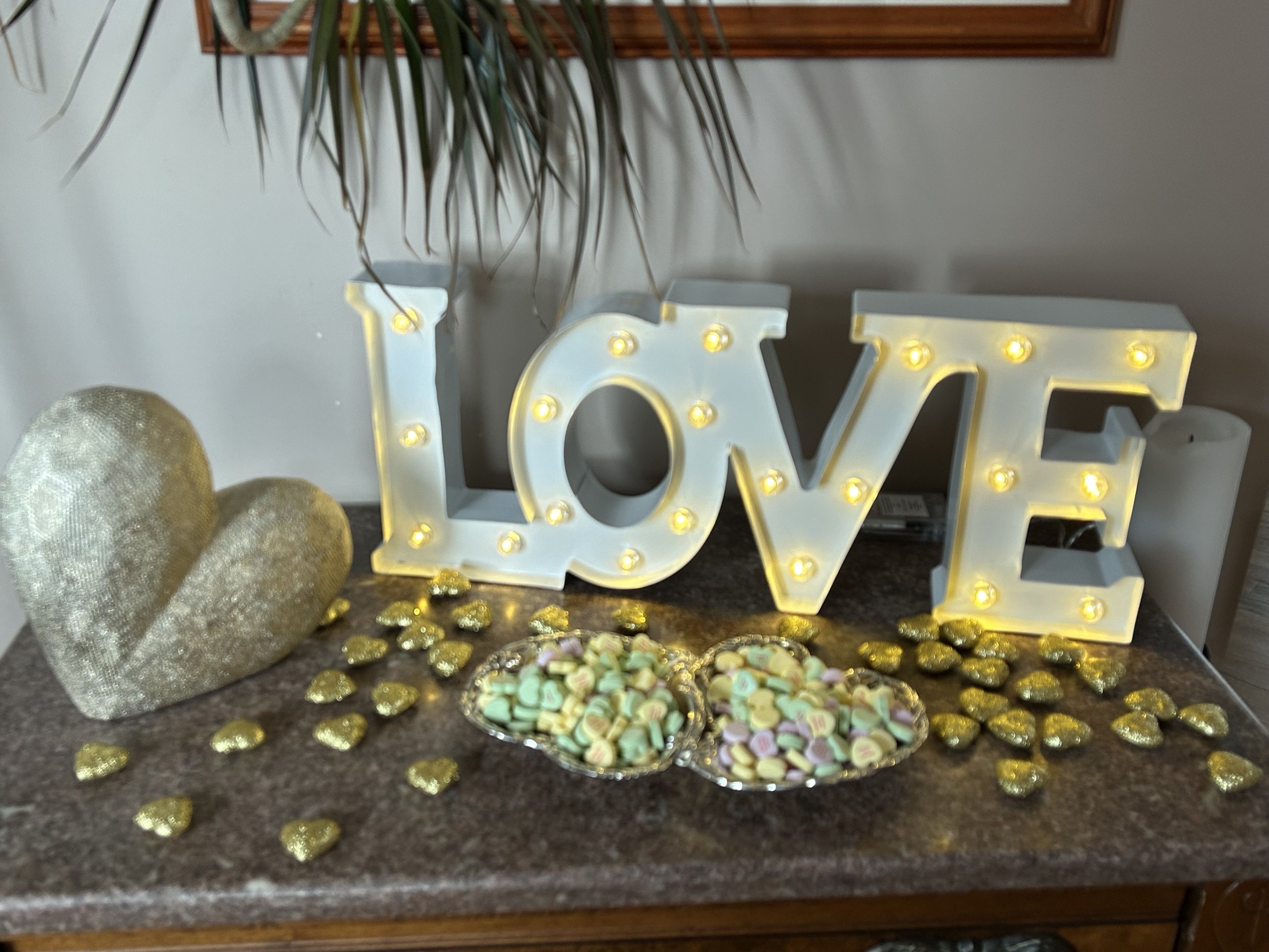 Decorative table arrangement with a large illuminated 'LOVE' sign, a gold glittery heart, two silver trays with pastel-colored candy hearts, and gold foil-wrapped chocolates on a brown marble surface.