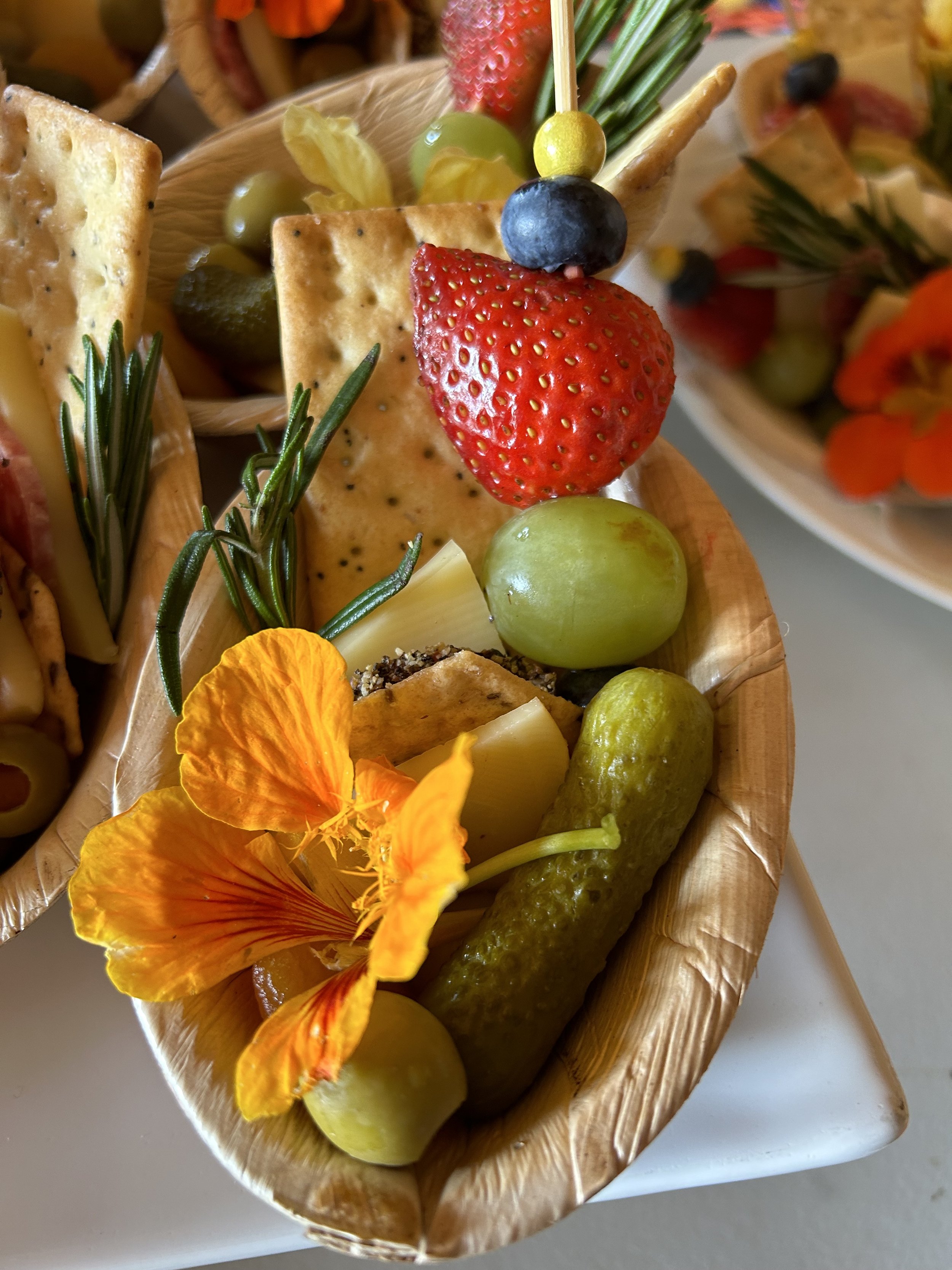 A basket contains fresh grapes, strawberries, crackers, a cucumber, a pickle, edible flowers, and sprigs of rosemary, arranged as a snack platter.