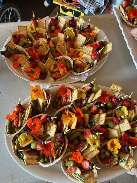 Two large platters filled with assorted cheese, crackers, fresh strawberries, grapes, and edible flowers, decorated with small skewers.