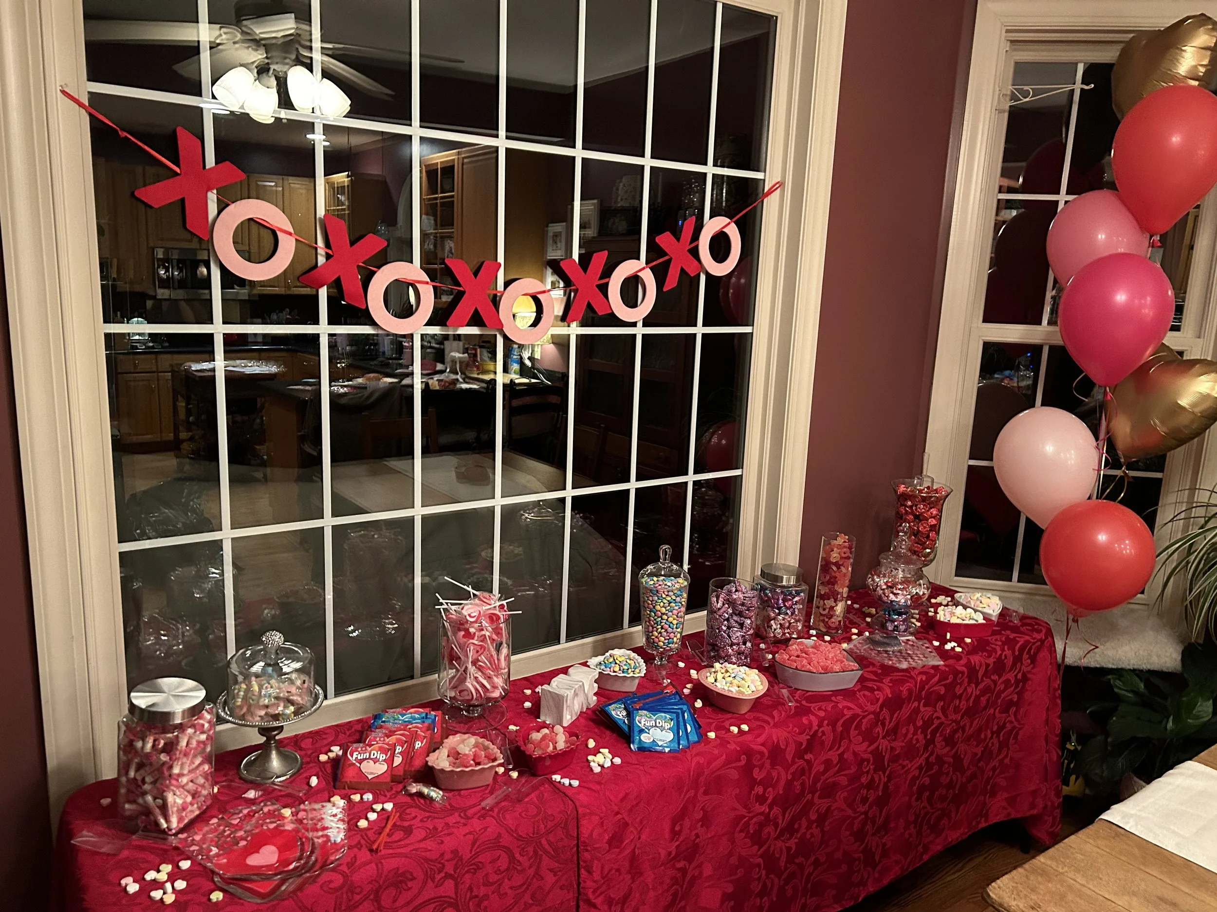 A Valentine's Day themed dessert table with a red tablecloth, assorted candies, and pink, red, and gold balloons, with a 'XOXO' banner hanging in front of a window.