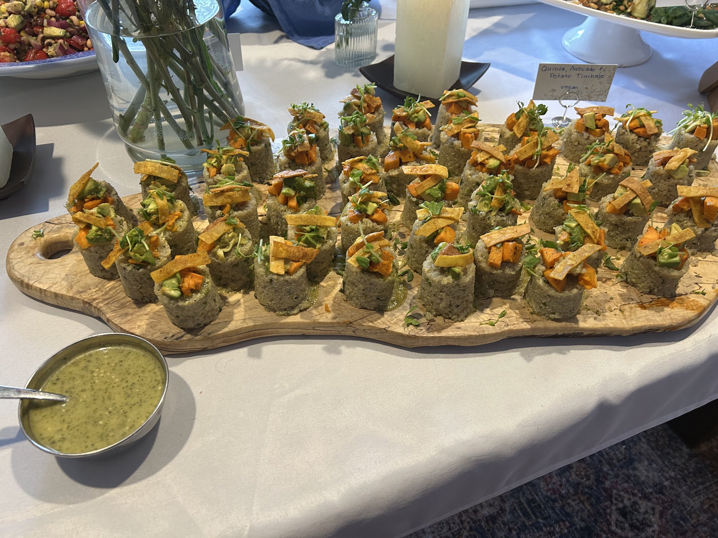 A wooden serving board with bite-sized vegan sushi topped with roasted vegetables and microgreens, accompanied by a small bowl of green dipping sauce, on a white tablecloth. In the background, there are salads and a candle.