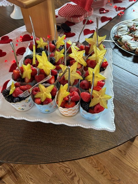 A tray of individual fruit cups topped with star-shaped pineapple slices and fruit skewers on a wooden table decorated for Valentine's Day.