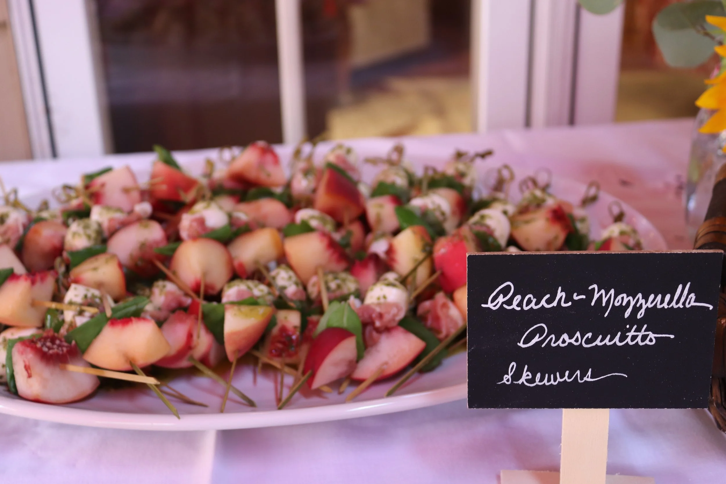 A platter of peach-mozzarella prosciutto skewers on a white tray with a black handwritten sign. The sign reads 'Peach-Mozzarella Prosciutto Skewers'.