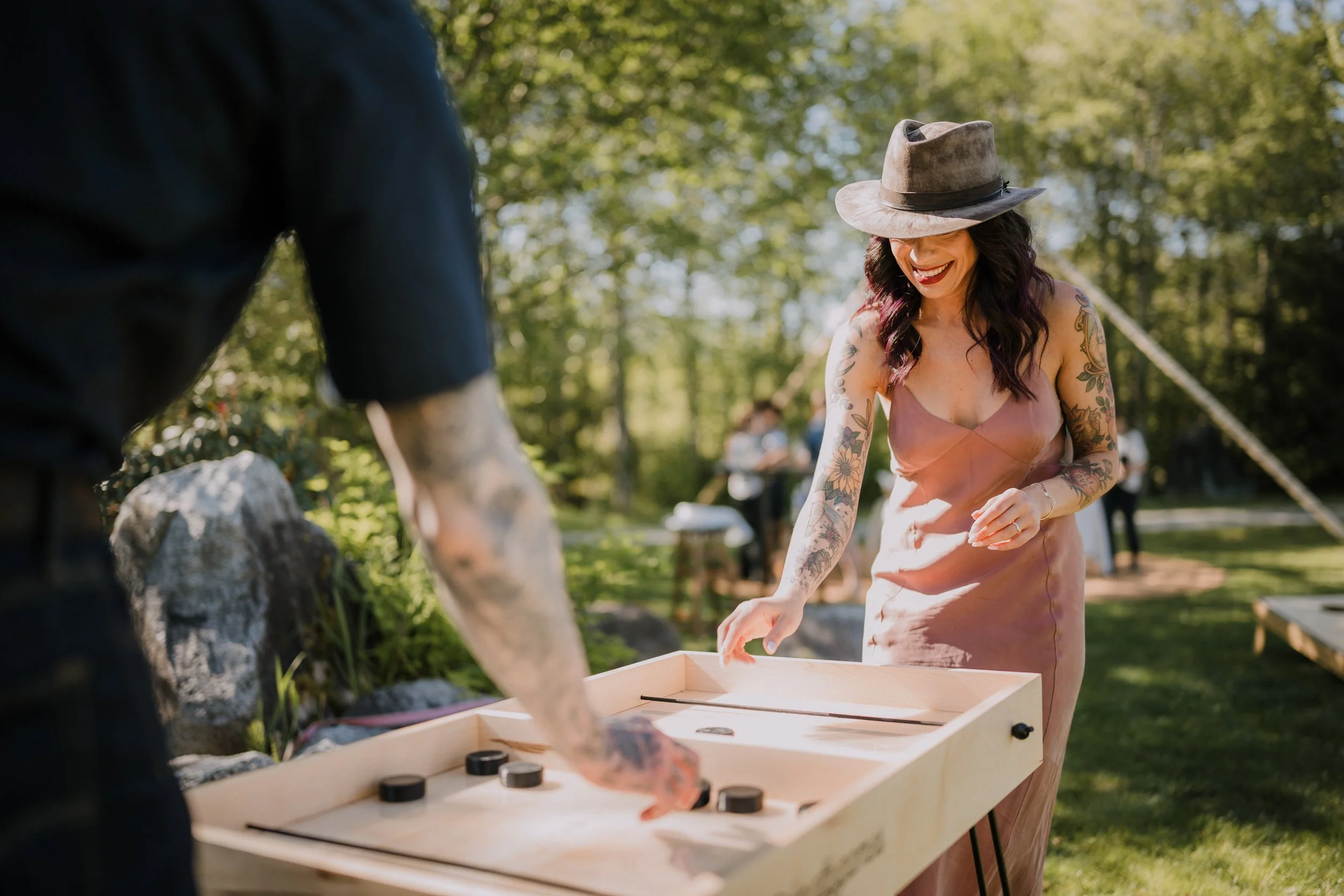 Two wedding guests with tattoos, one off screen and one wearing a dress and a hat, play pucket