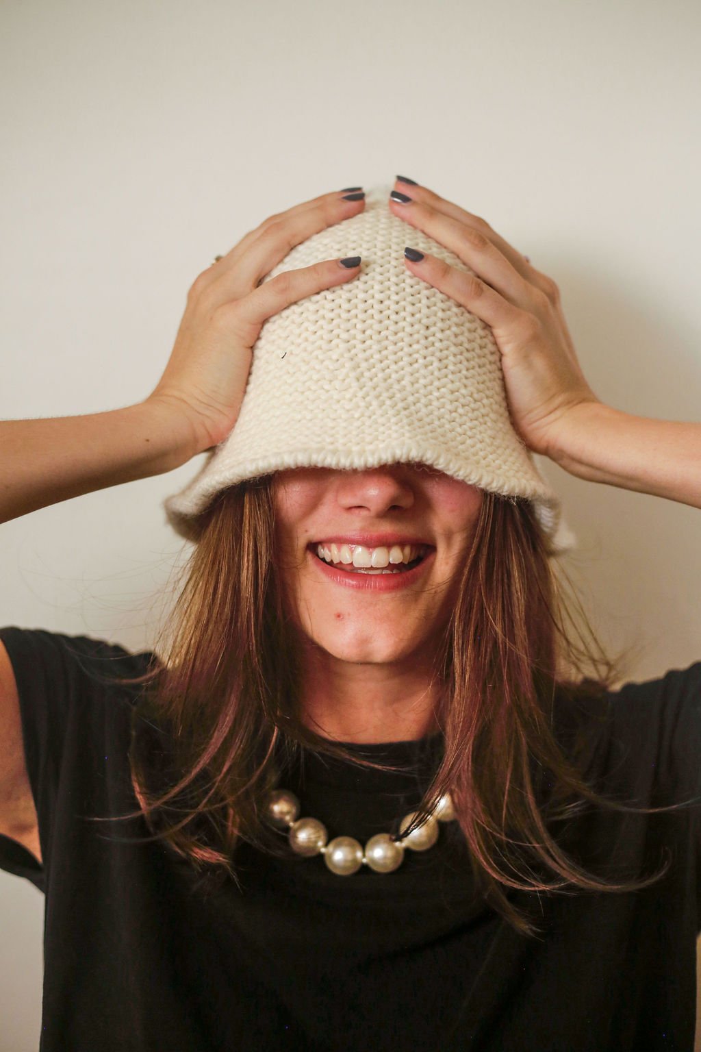A woman with long brown hair, smiling with her teeth showing, wearing a black shirt and a large pearl necklace, is holding a white knit hat over her eyes with both hands.