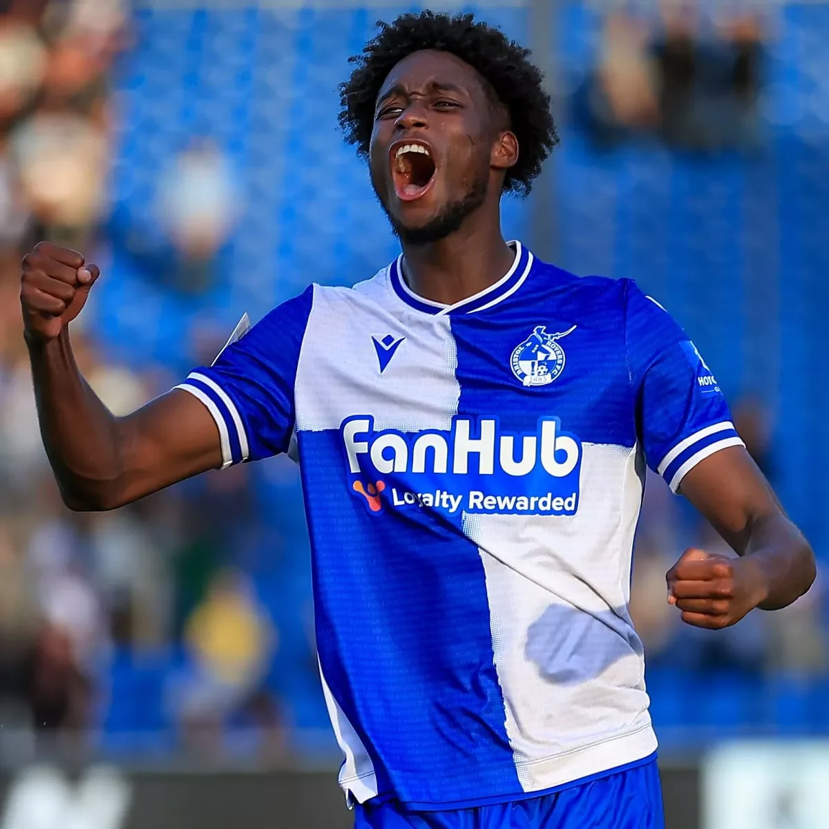 A soccer player in a blue and white jersey celebrating on the field with a clenched fist and open mouth.