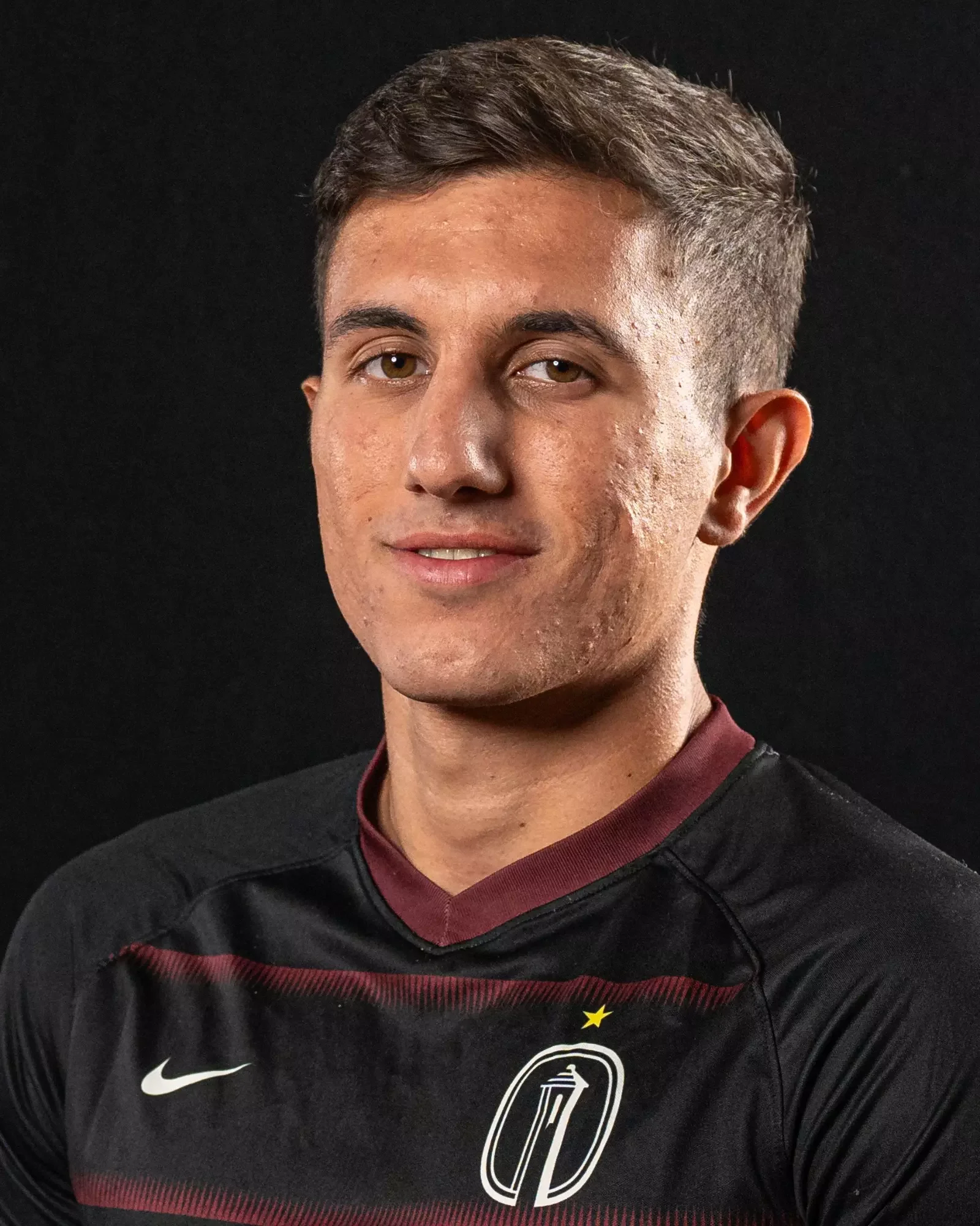 Portrait of a young man wearing a black sports jersey with a logo and Nike swoosh, against a black background.
