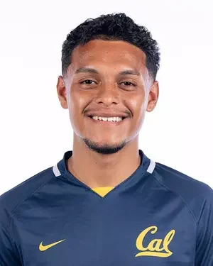 A young man with curly black hair and a beard, smiling, wearing a dark blue sports jersey with yellow accents and the word 'Cal' on it.